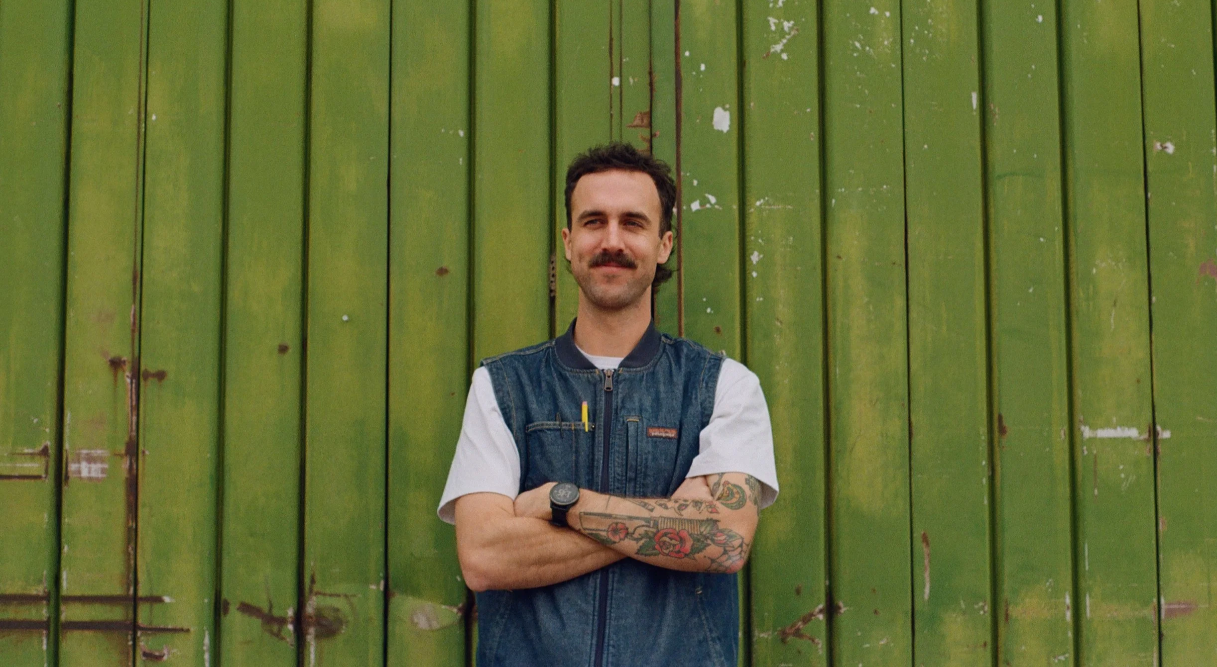 A man with dark hair, a mustache, and tattoos on his arm, wearing a denim vest over a white t-shirt, stands in front of a green wooden wall with his arms crossed and a slight smile.