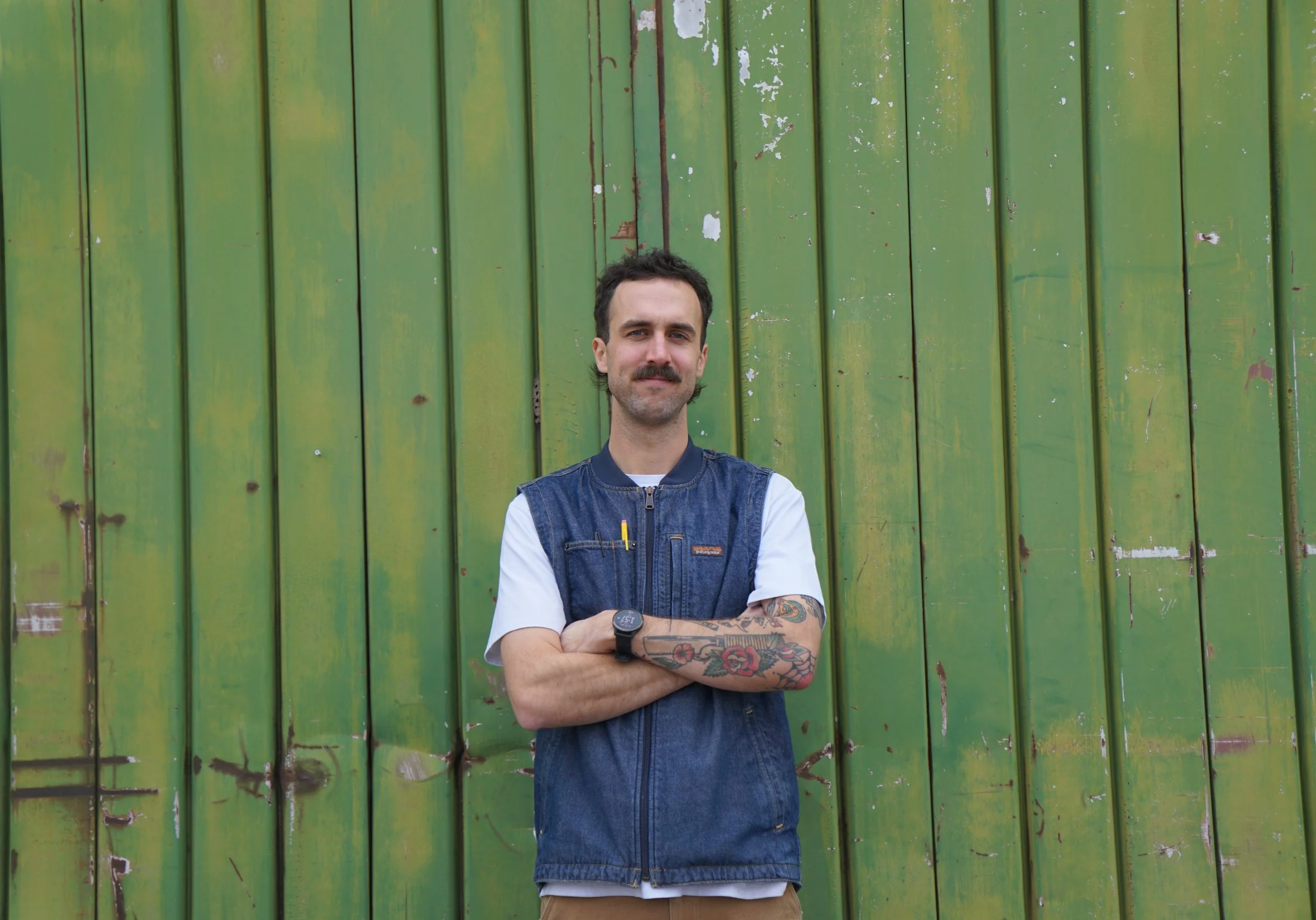 Ethan Miller with, owner and operator of Granary Haus, a custom furniture studio in Salt Lake City, Utah, with arms crossed in front of a green wall