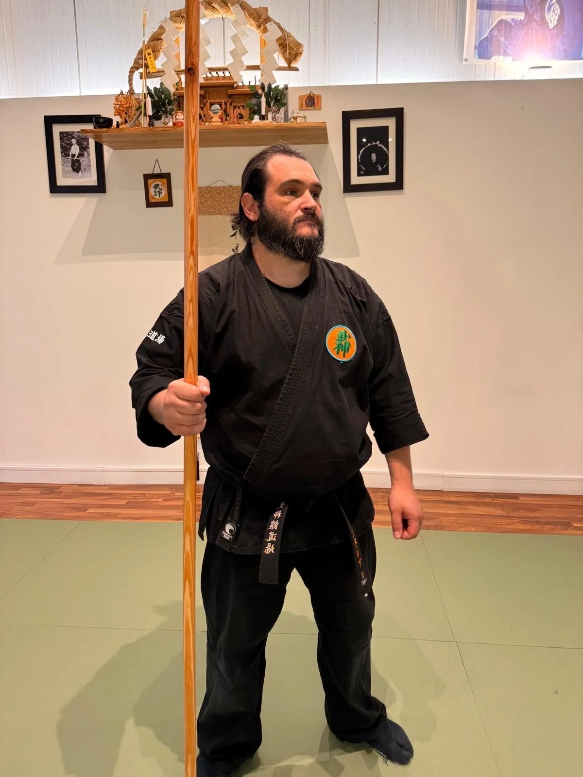 Andrea Traversa in uniforme bujinkan con cintura nera, che tiene in mano un'arma lunga in legno, in un dojo, con decorazioni giapponesi e fotografie sul muro.