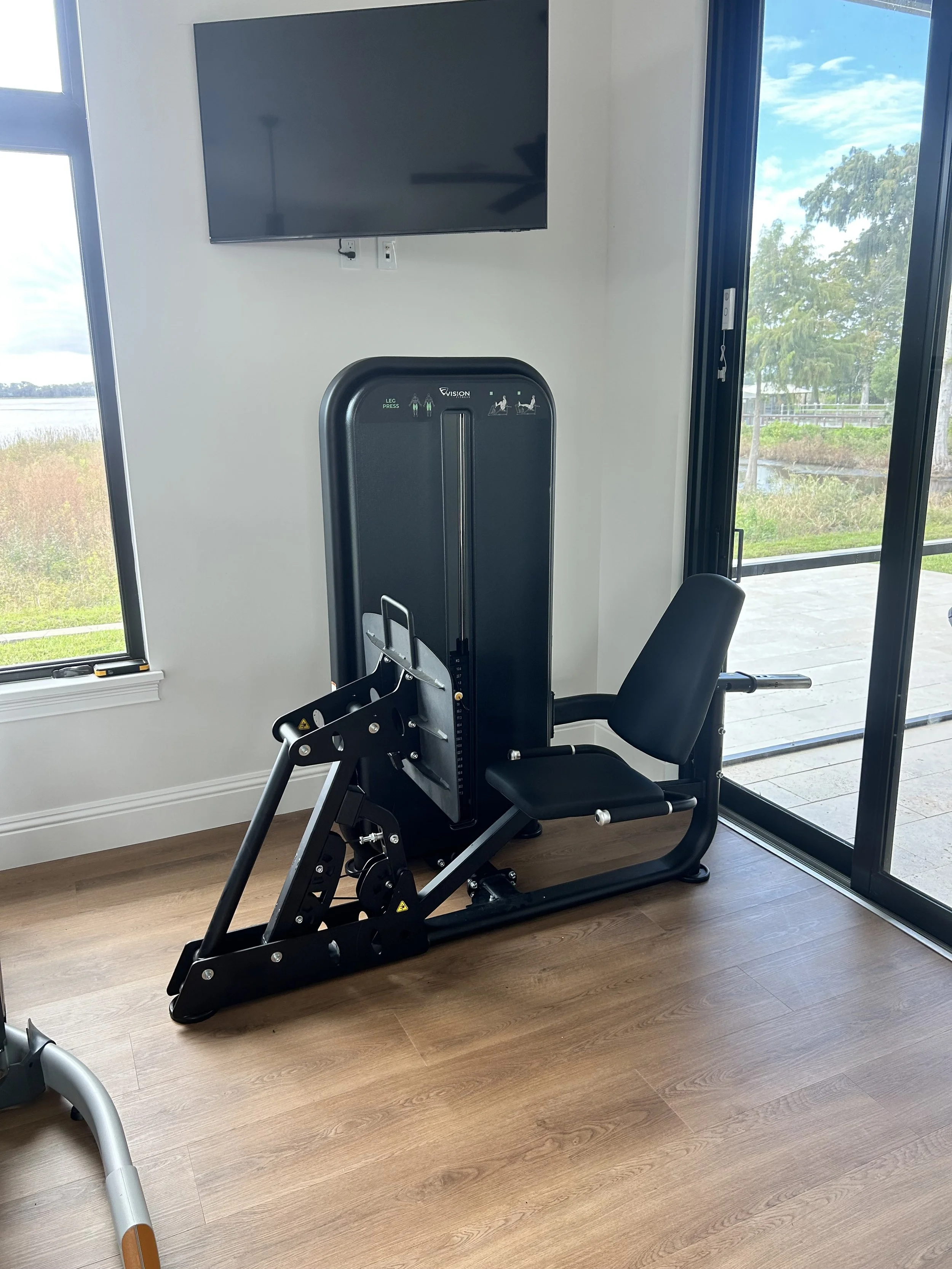 Seated exercise machine with a padded seat and backrest, positioned near large windows and a sliding glass door with an outdoor view of grass and trees.