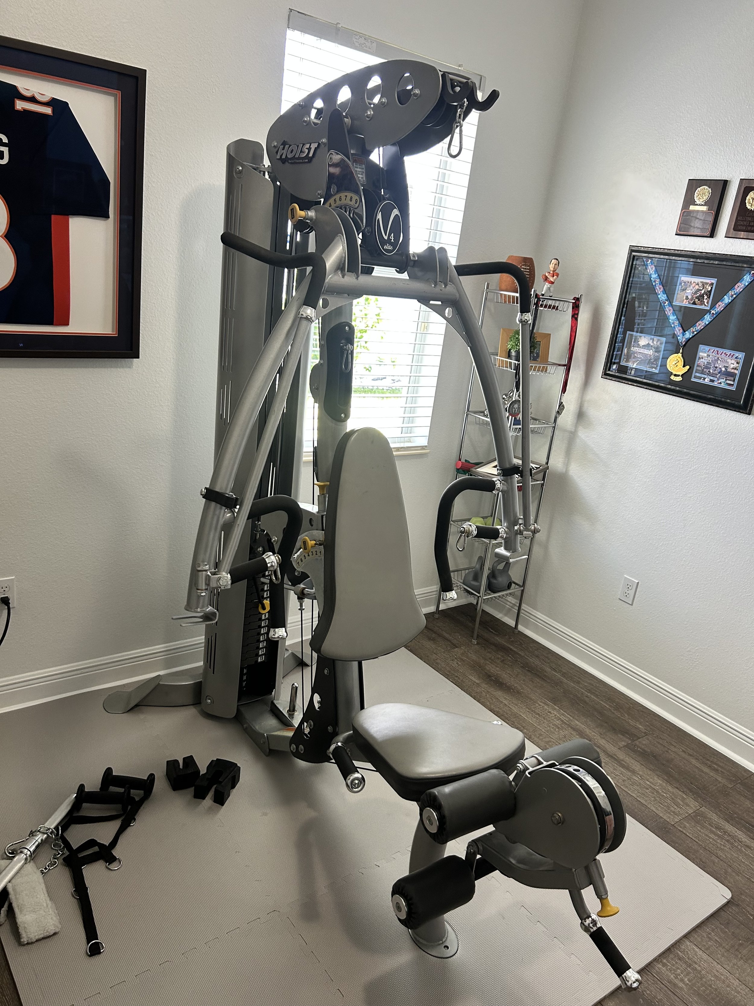 Home gym equipment with a multi-function weight machine, resistance bands, and a small step platform, situated in a room with framed jerseys and medals on the wall.