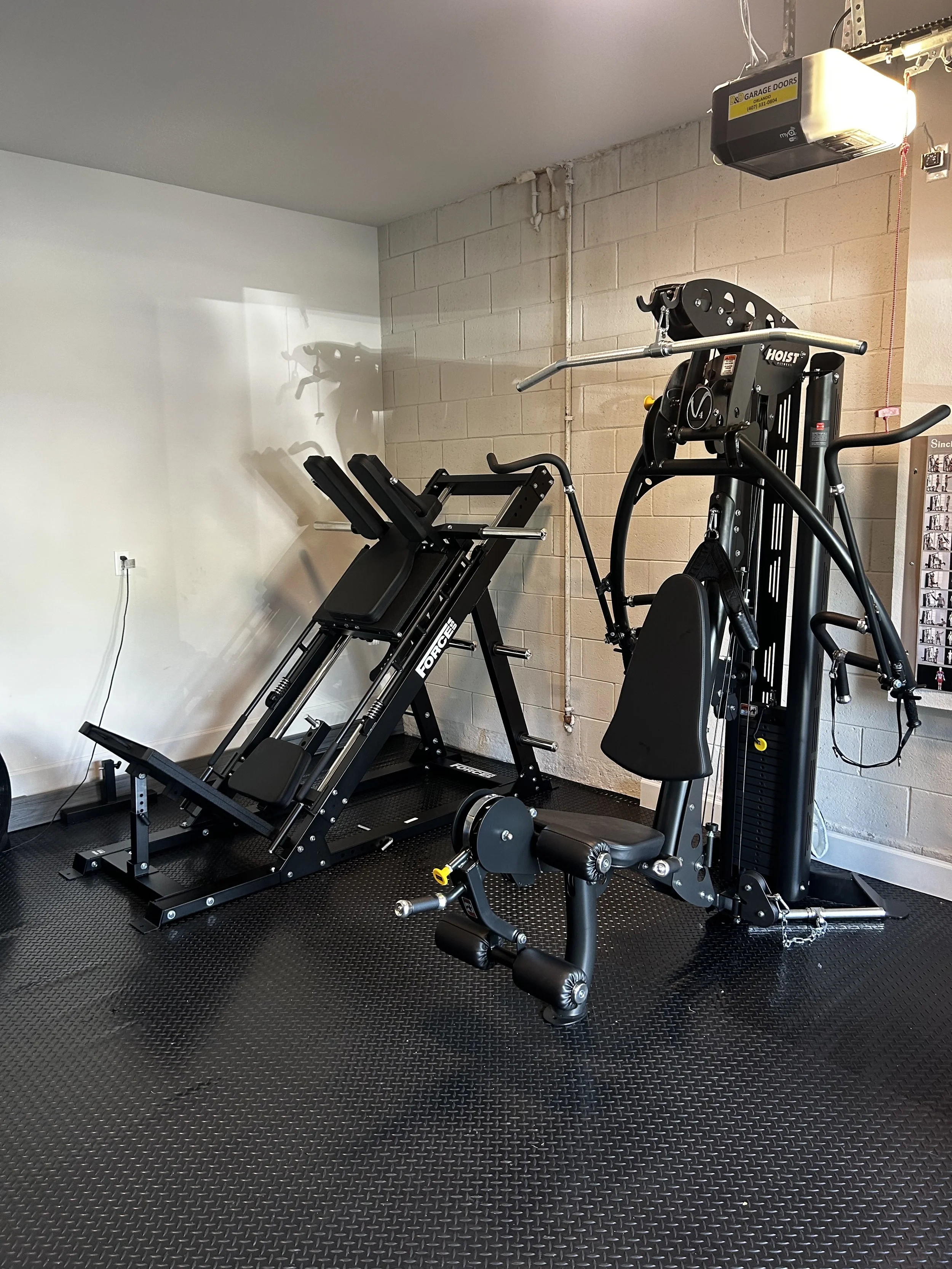 Garage gym with a black exercise bike and a multi-station workout machine on a black rubber floor, against a cinder block wall.