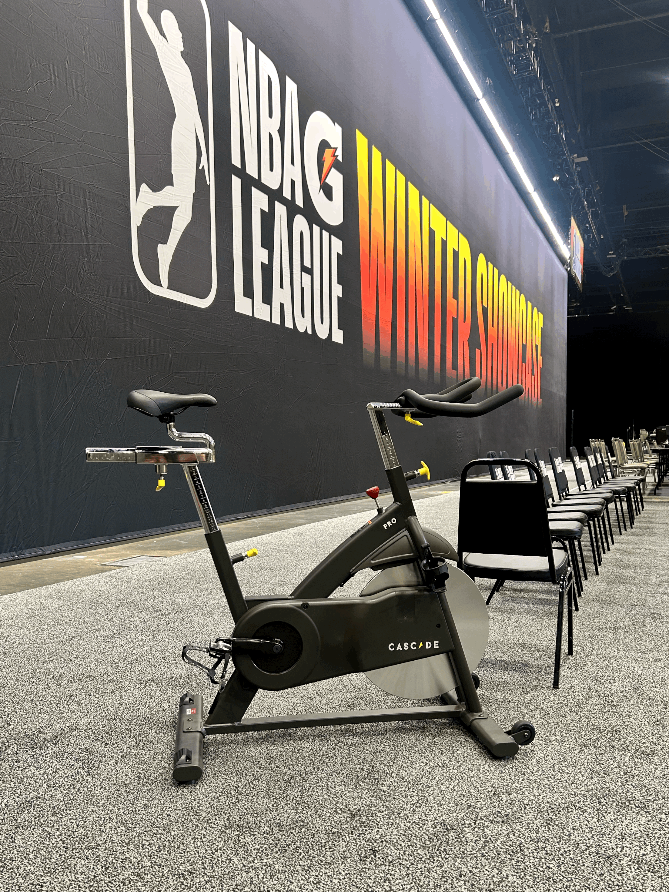 Indoor scene with a stationary exercise bike in front of a NBA G League Sign with a row of black chairs along the side.