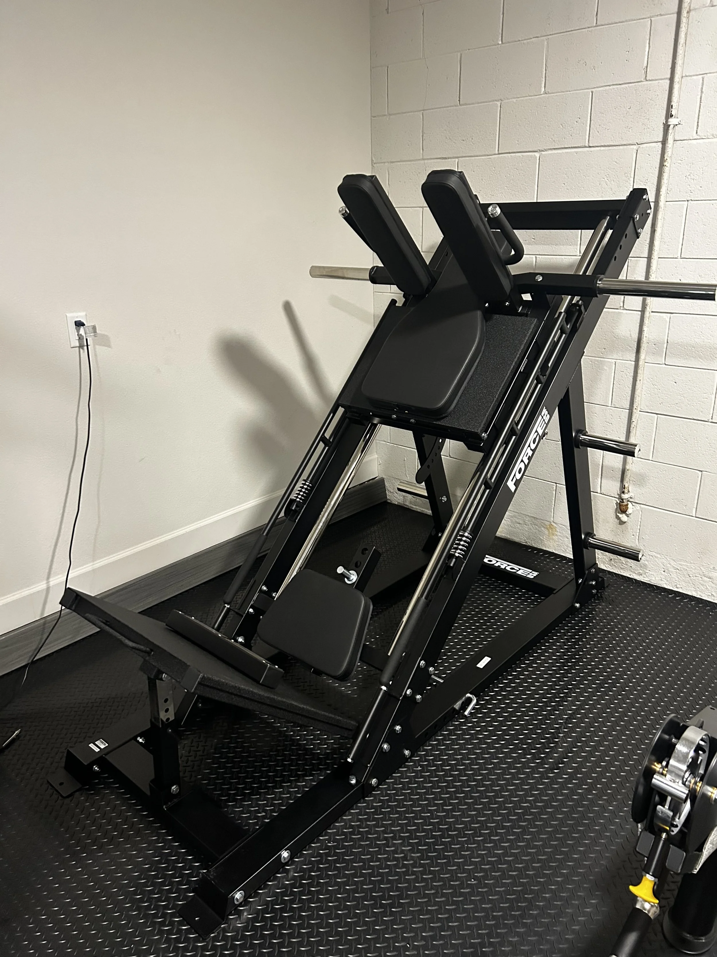 Powerlifting squat rack and stand with safety bars, black padded supports, and weight storage pegs, placed on a black rubber mat in a gym room