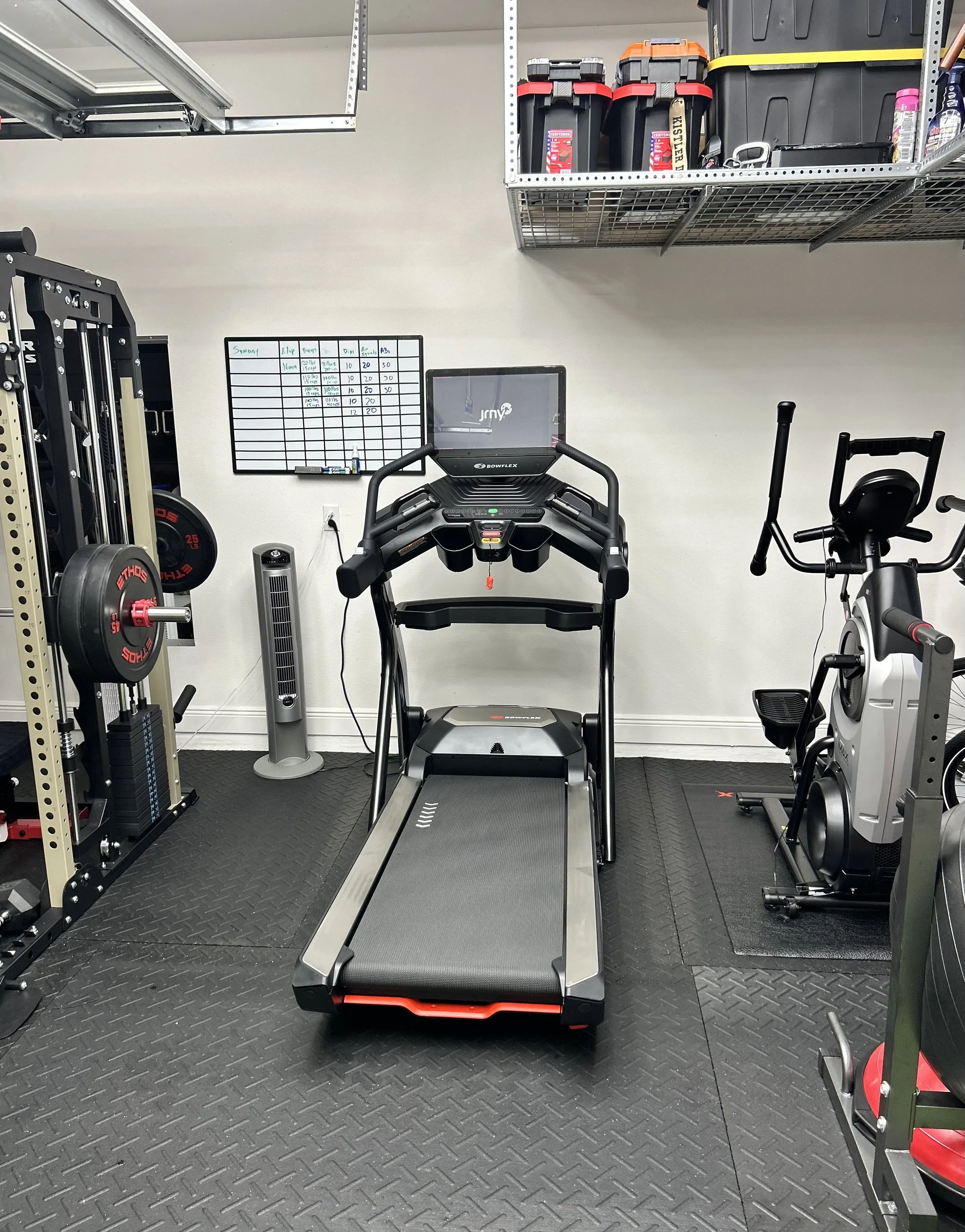 A home gym with a treadmill, exercise bike, weight rack, and shelving unit with storage containers.