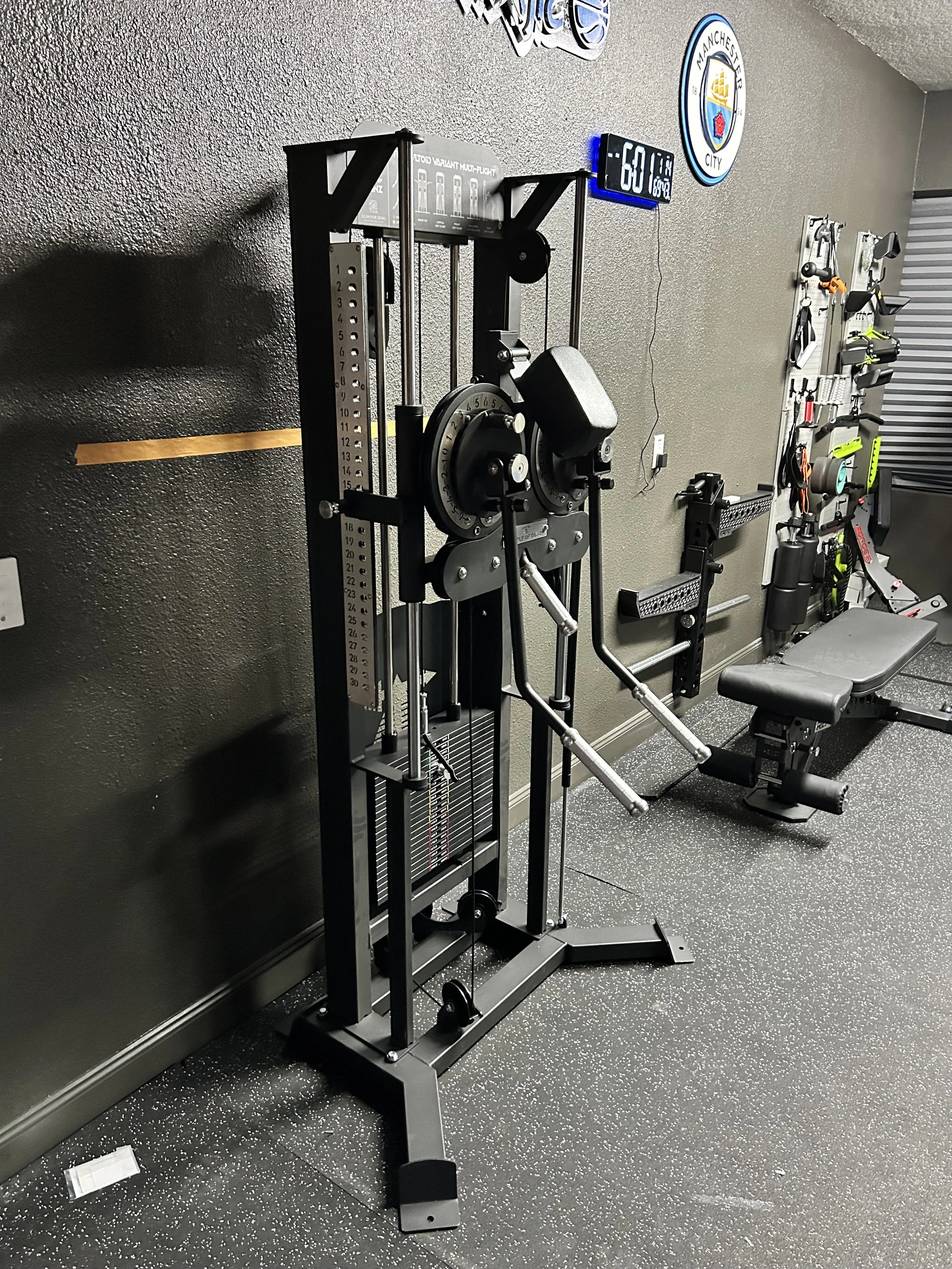 A fitness gym corner with a black chest press machine, an adjustable weight bench, and various workout equipment on a wall-mounted rack. A digital clock displaying 6:50 is on the wall.