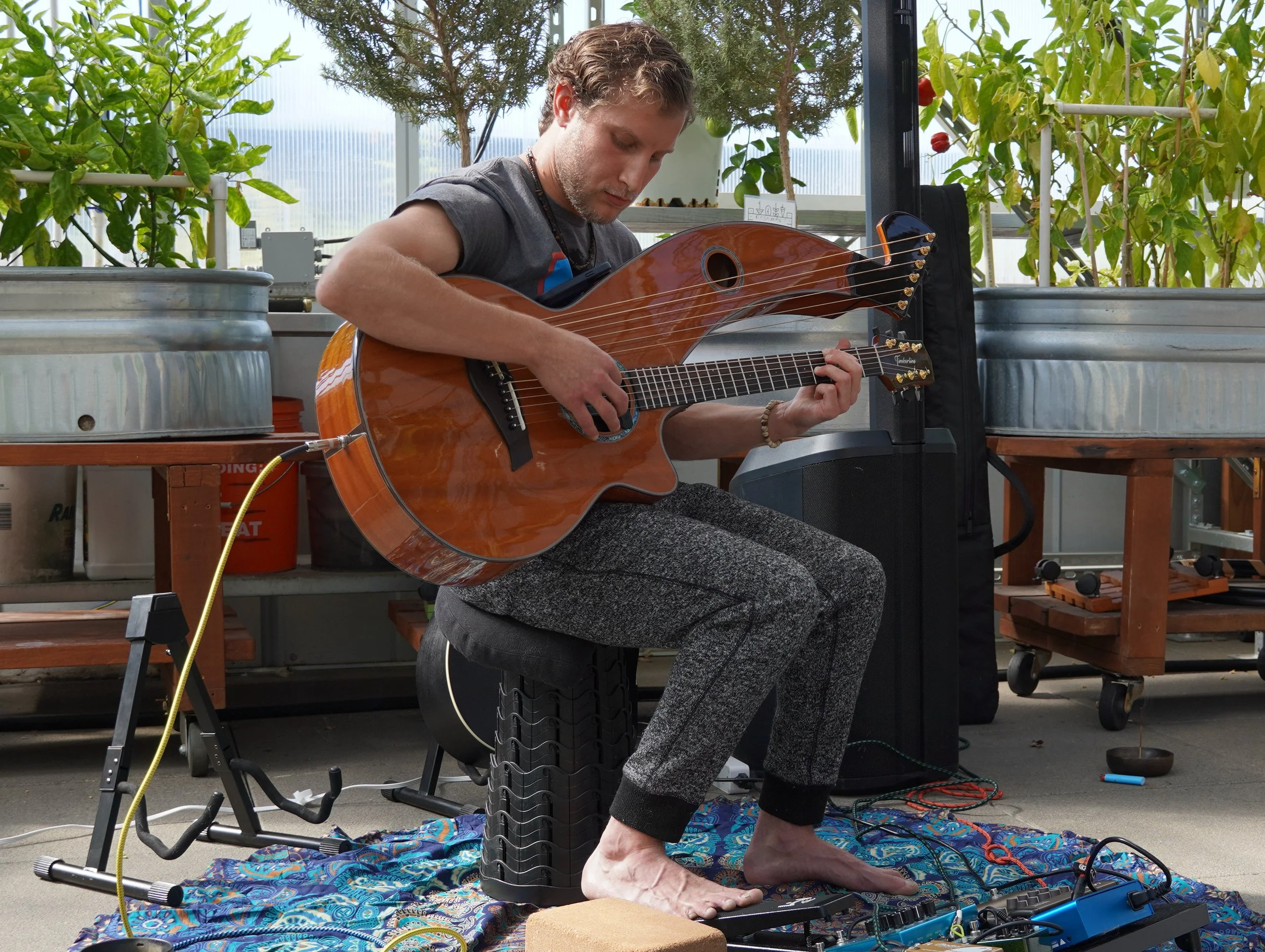 A man playing an acoustic guitar while sitting on a stool in a greenhouse or garden area. He's barefoot, with various equipment and plants around him.