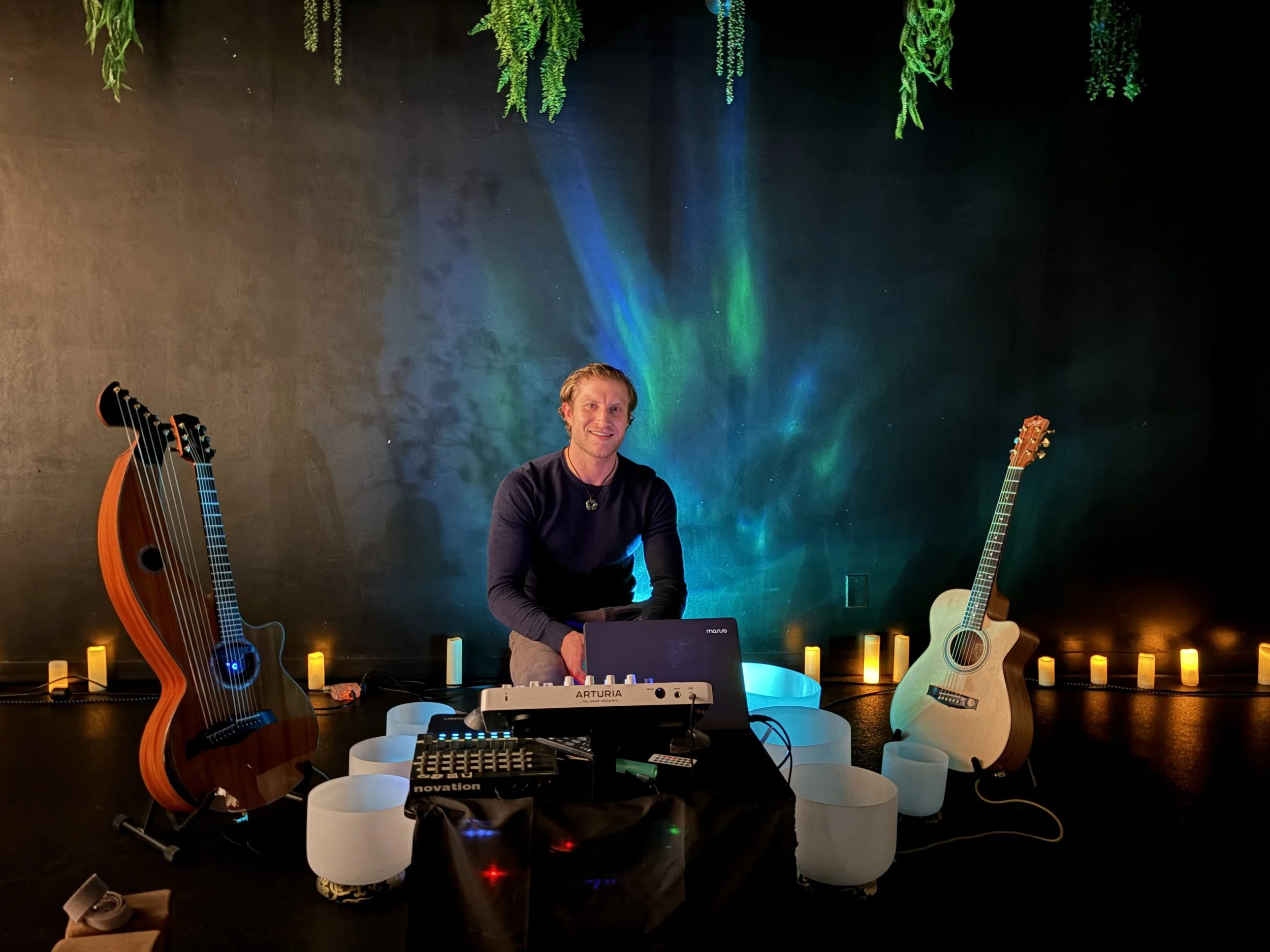 A man sitting behind a music DJ setup with two guitars, one electric and one acoustic, in a room with dark walls, colorful herbs hanging from the ceiling, and small lit candles along the back wall.