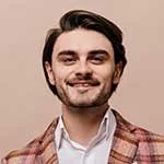 A young man with dark hair and a beard, wearing a plaid blazer and white shirt, smiling at the camera