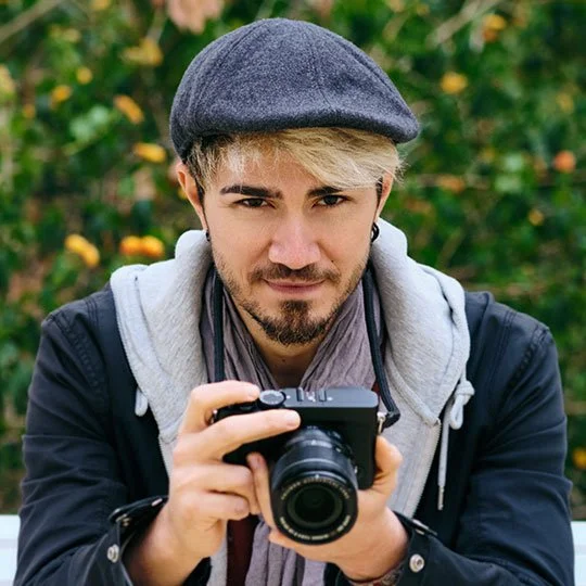 Young man with blond hair and beard, wearing a gray cap, black jacket, and gray hoodie, holding a camera outdoors with a blurred green and yellow background.