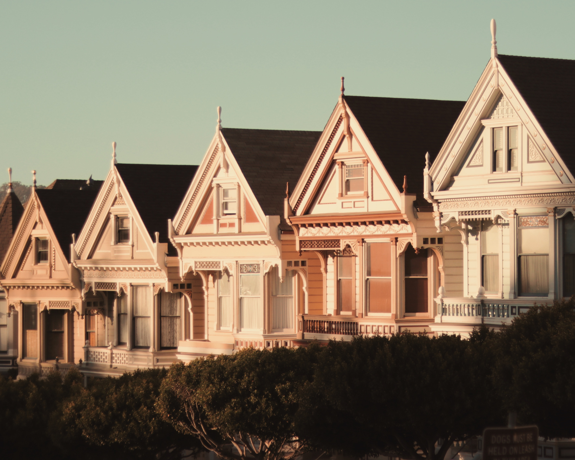 A row of Victorian-style houses with ornate detailing and front porches, set against a clear sky, with trees in the foreground.