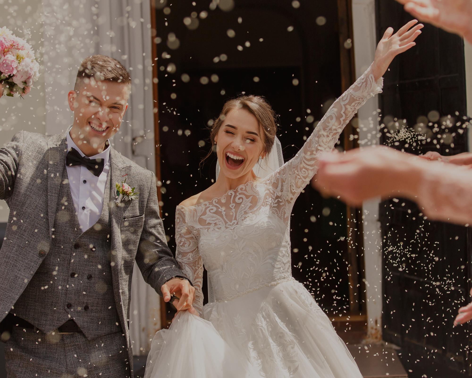 A newlywed couple celebrating as they walk through confetti and rice, smiling and holding hands outside a building, during their wedding ceremony.