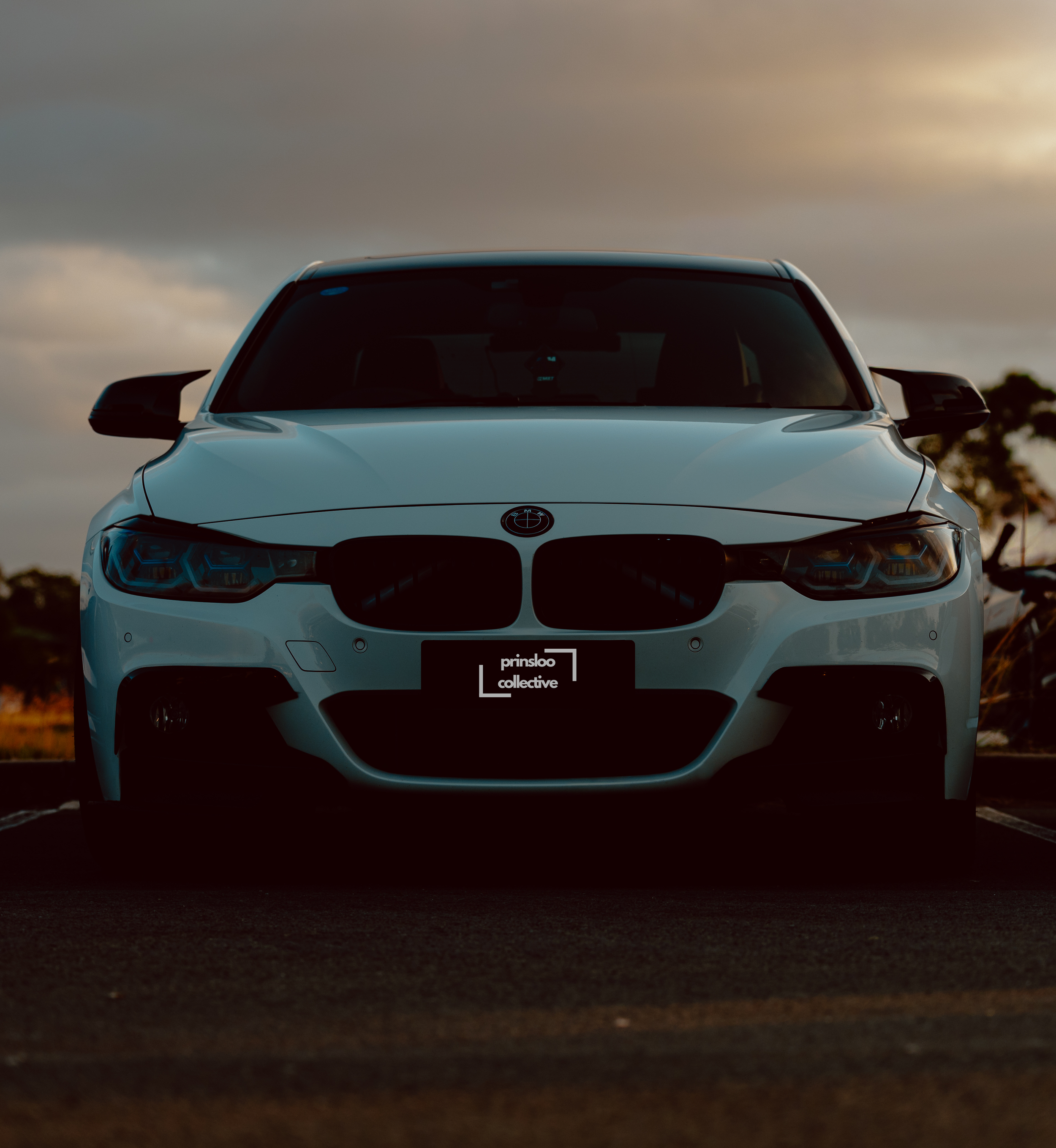 Front view of a white BMW car parked outdoors during sunset, with a dark sky and trees in the background.