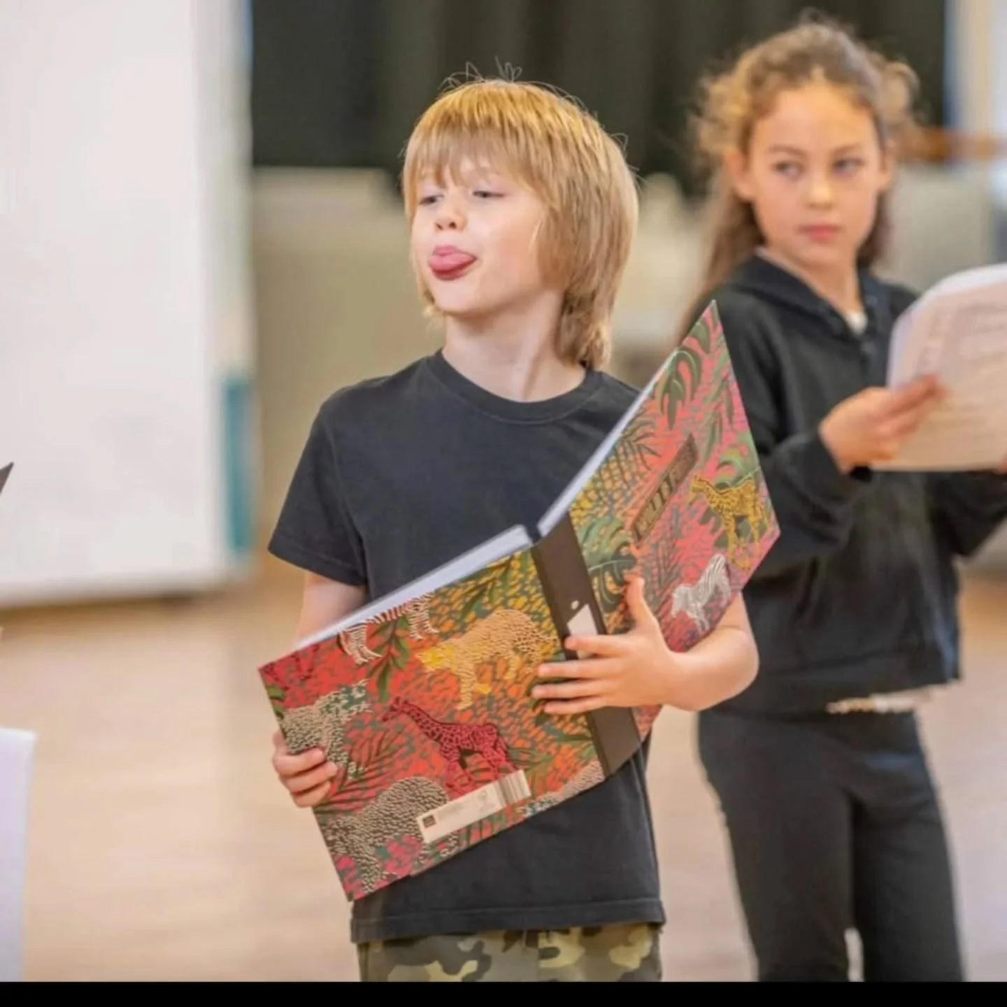 A young boy with red hair wearing a black t-shirt makes a funny face while holding a colorful sketchbook. A girl with curly hair and a black hoodie stands in the background holding a notepad, in what appears to be a classroom or art studio.