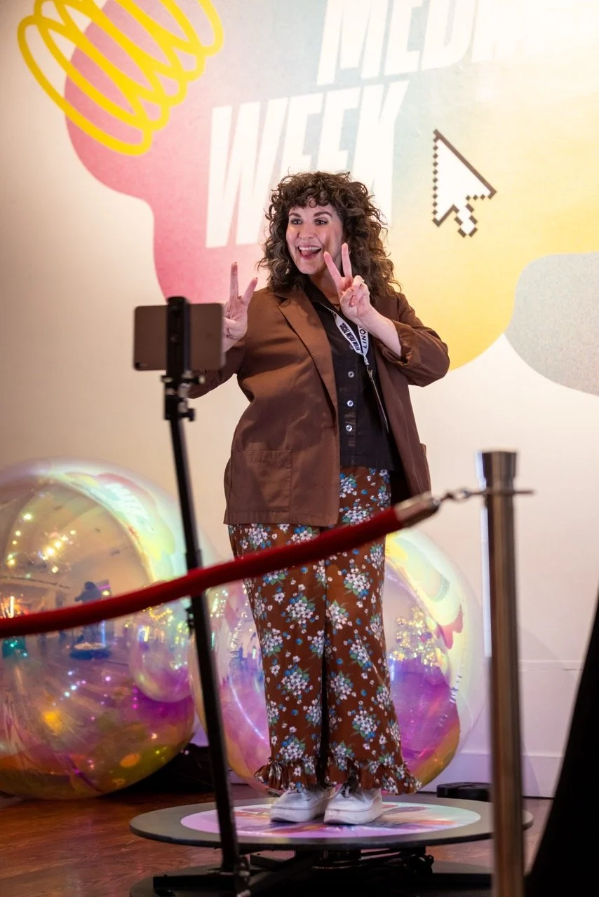 A woman with curly hair taking a selfie, making peace signs with both hands, standing on a platform decorated with gold and purple balloons, with a colorful background that includes a large pink and yellow graphic and white computer mouse pointer icon.