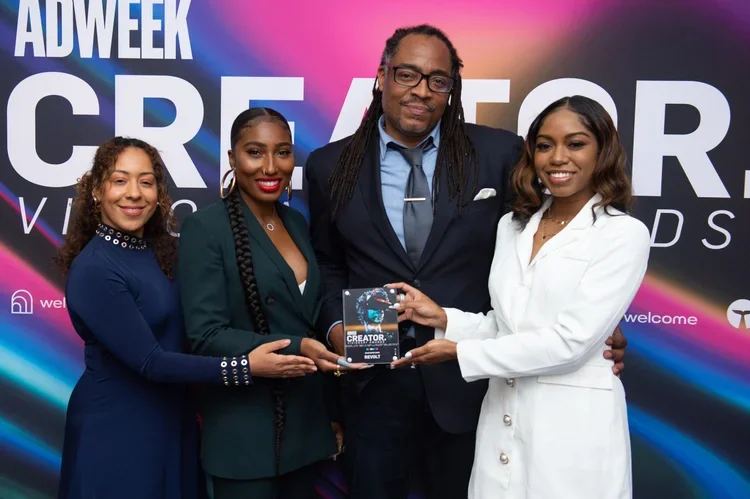 Four individuals standing together at an event, three women and one man, holding an award plaque. The backdrop features large text reading 'ADWEEK CREATOR' and colorful graphics.