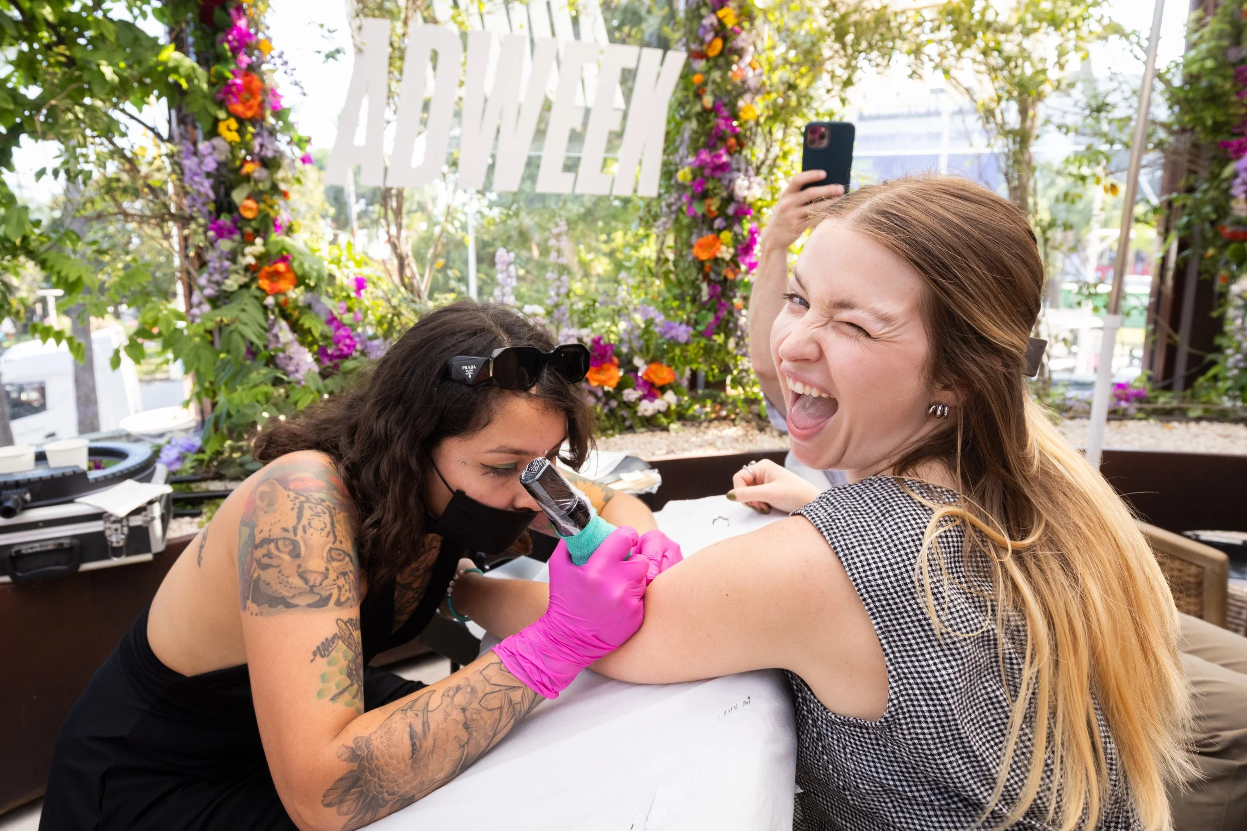 A woman gets a tattoo on her upper arm by a tattoo artist in an outdoor setting decorated with flowers, with a large 'FLOWER' sign in the background. The woman is laughing and winking while the artist is focused on her work.