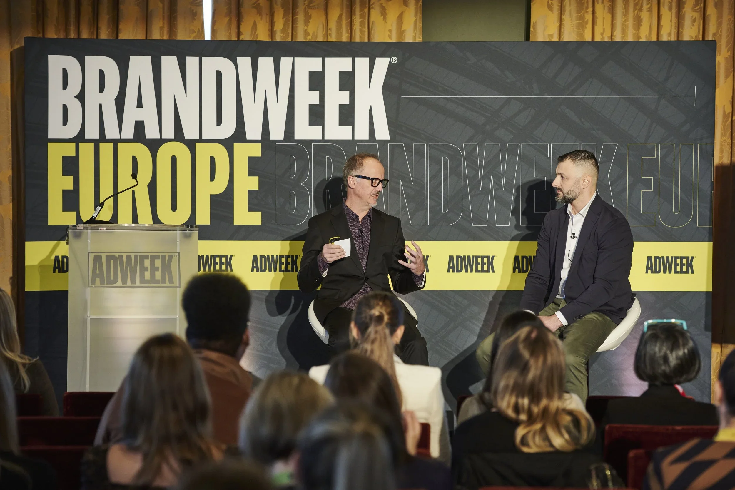 Two men in conversation on stage at a conference, with an audience in front. The backdrop reads "BRANDWEEK EUROPE" and "ADWEEK" in large text.