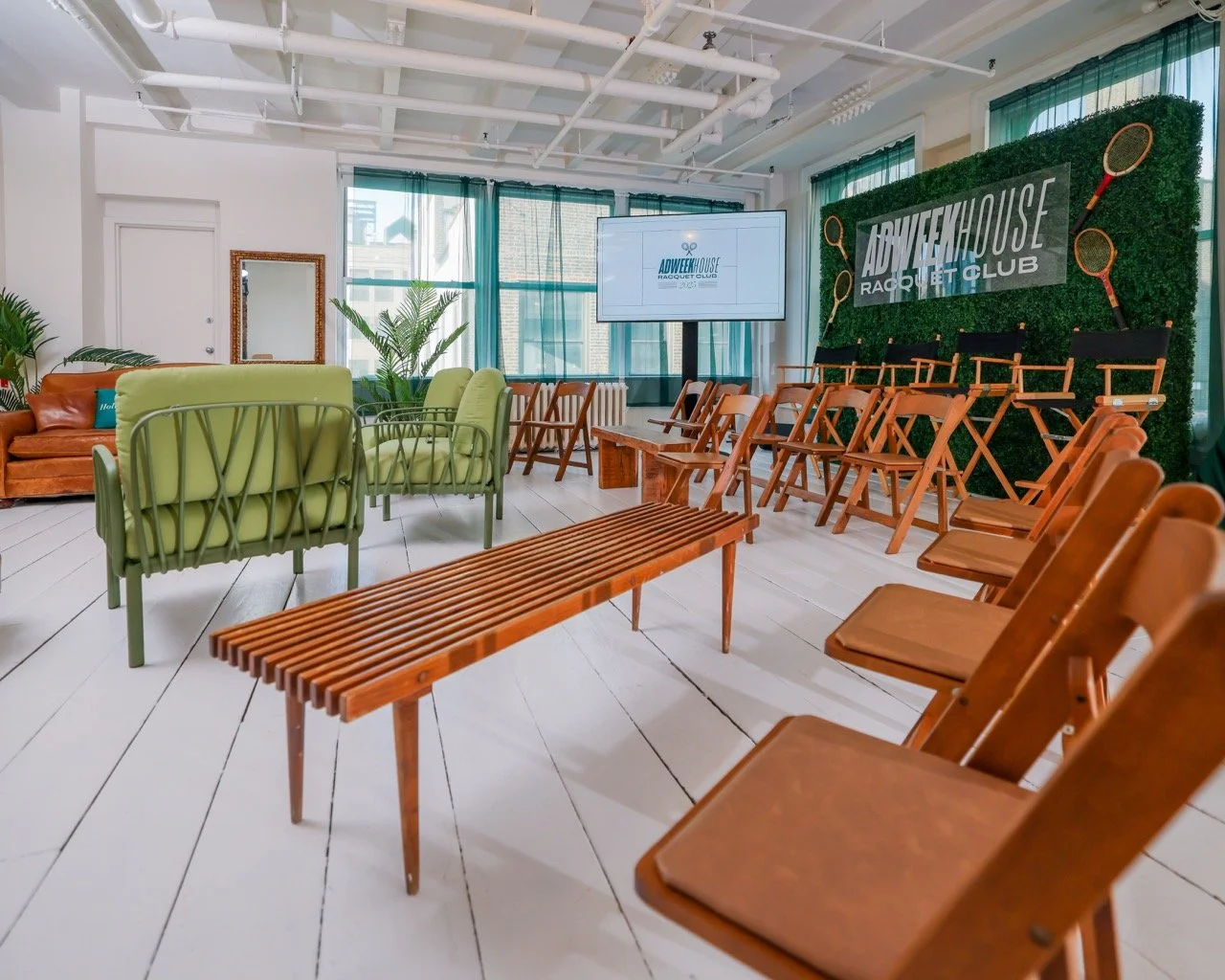Indoor space set up for an event with stacked chairs, a sofa, and a green backdrop with tennis rackets, featuring a large screen displaying 'ADWEKHOUSE RACQUET CLUB' in a bright, modern room with large windows and white floors.