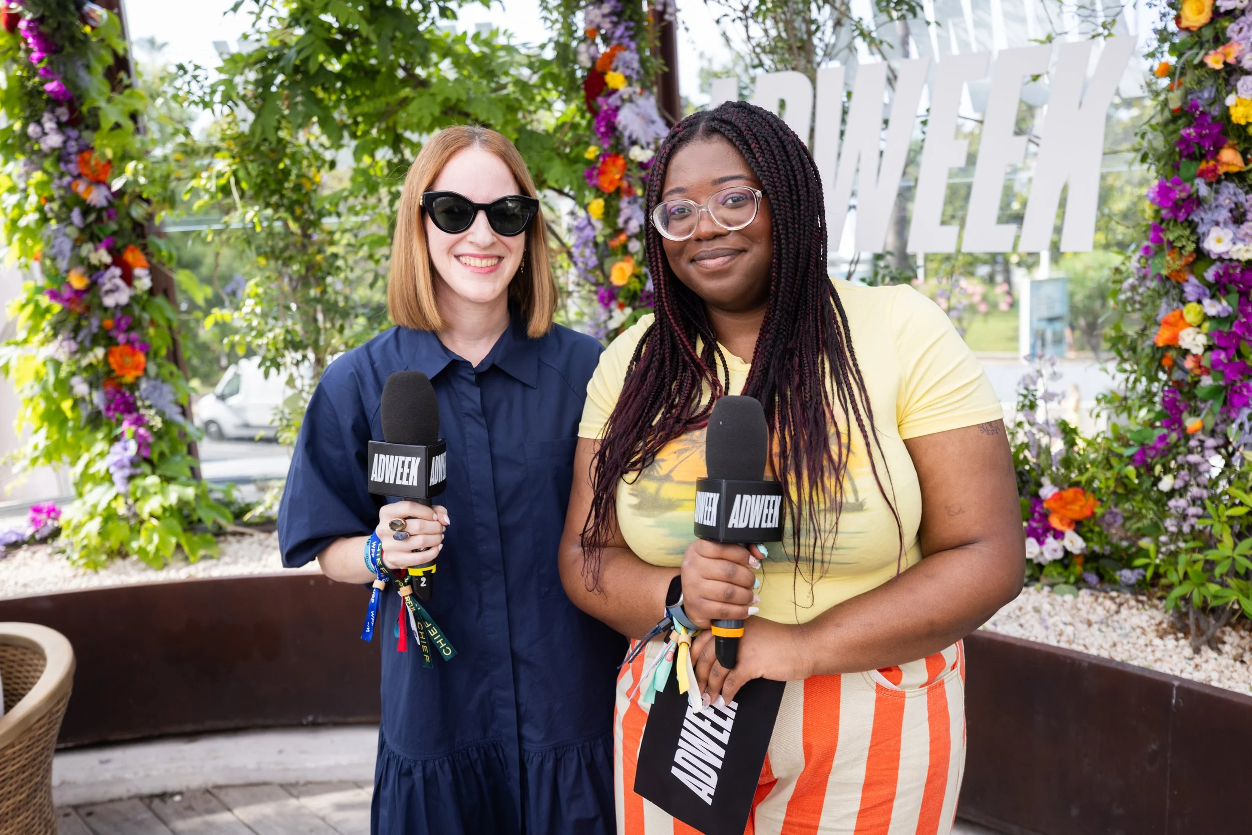 Two women holding microphones with ADWEEK logos, standing in front of a floral decoration at an outdoor event.
