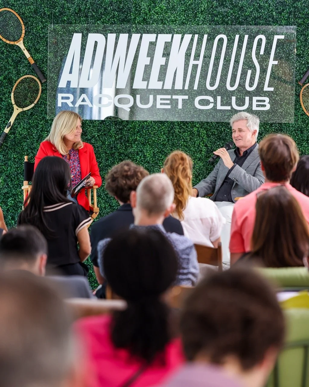 A panel discussion at the Avenuehouse Racquet Club with a woman in a red blazer and a man in a gray jacket speaking to an audience, surrounded by tennis racquets and a large sign reading 'AVENUEHOUSE RACQUET CLUB' on a green textured background.