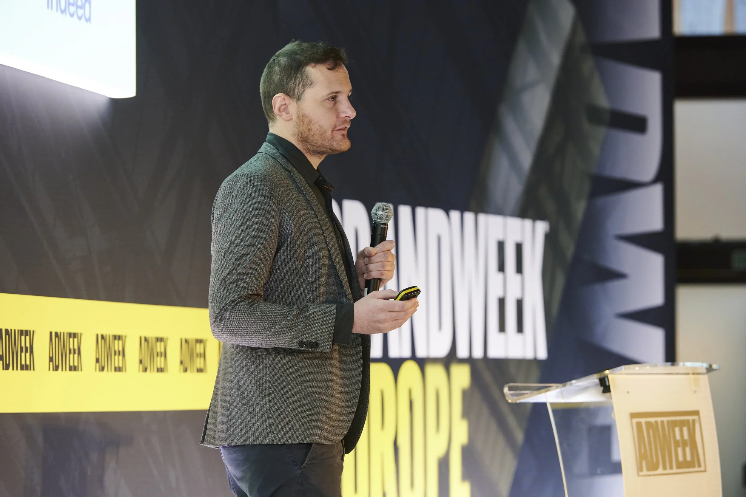 Man in gray blazer holding a microphone and a device, standing on stage at a conference or event.