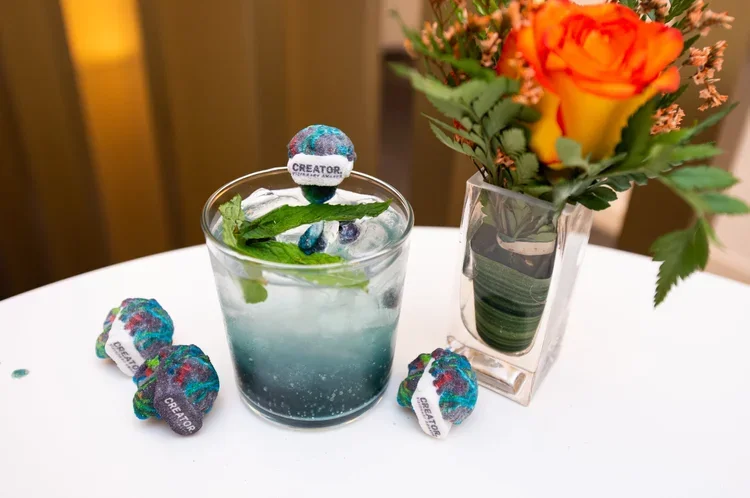 A white table with a clear glass of water with a small foam ball labeled 'Creator' on top, surrounded by four multicolored fabric balls, and a clear glass vase with orange and yellow roses and greenery.