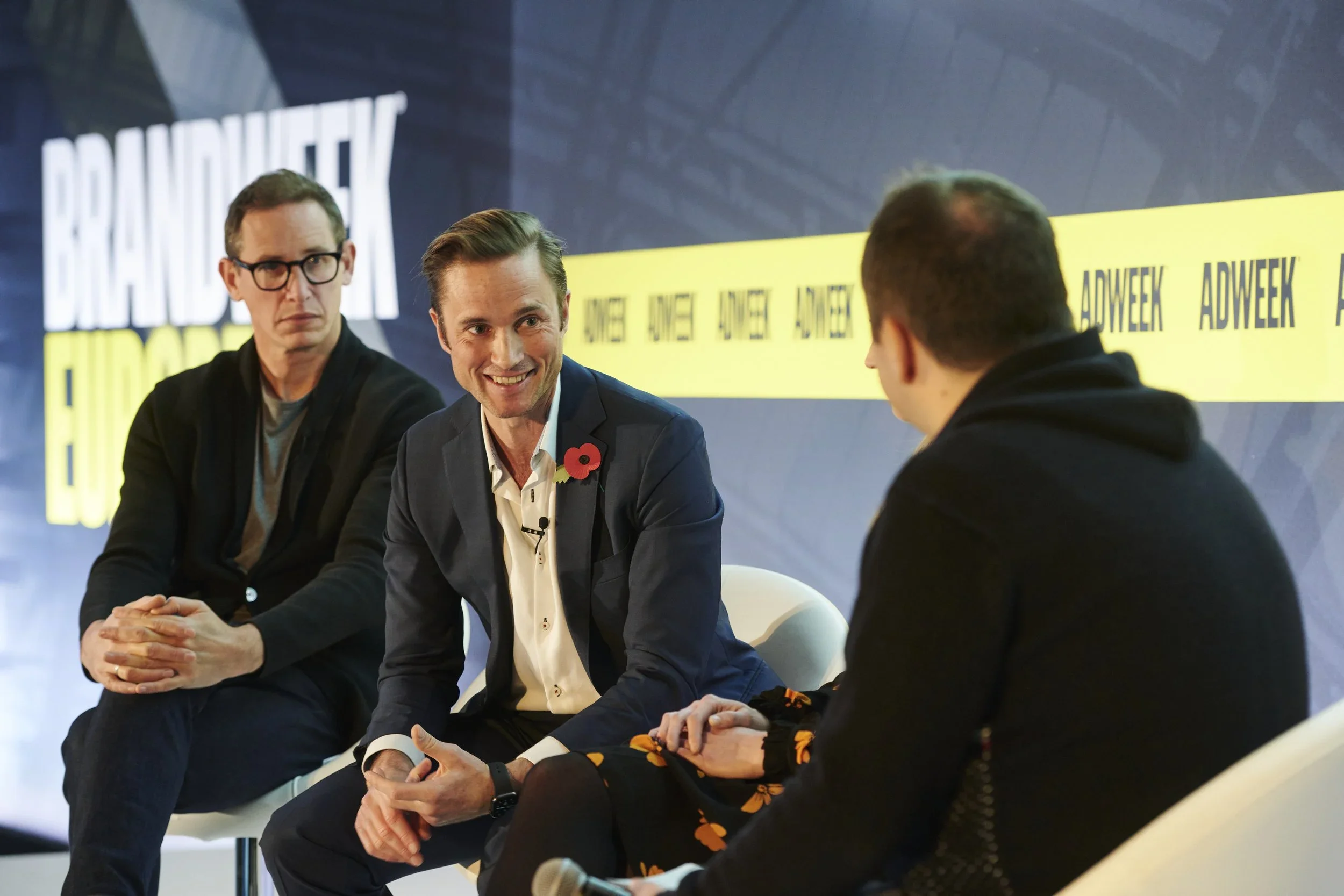 Three men sitting on a stage in conversation, with a large screen behind them displaying 'BRAND WEEK.' The man in the middle is smiling and wearing a suit with a poppy pin. The man on the left has glasses and a serious expression, and the man on the right is seen from behind.
