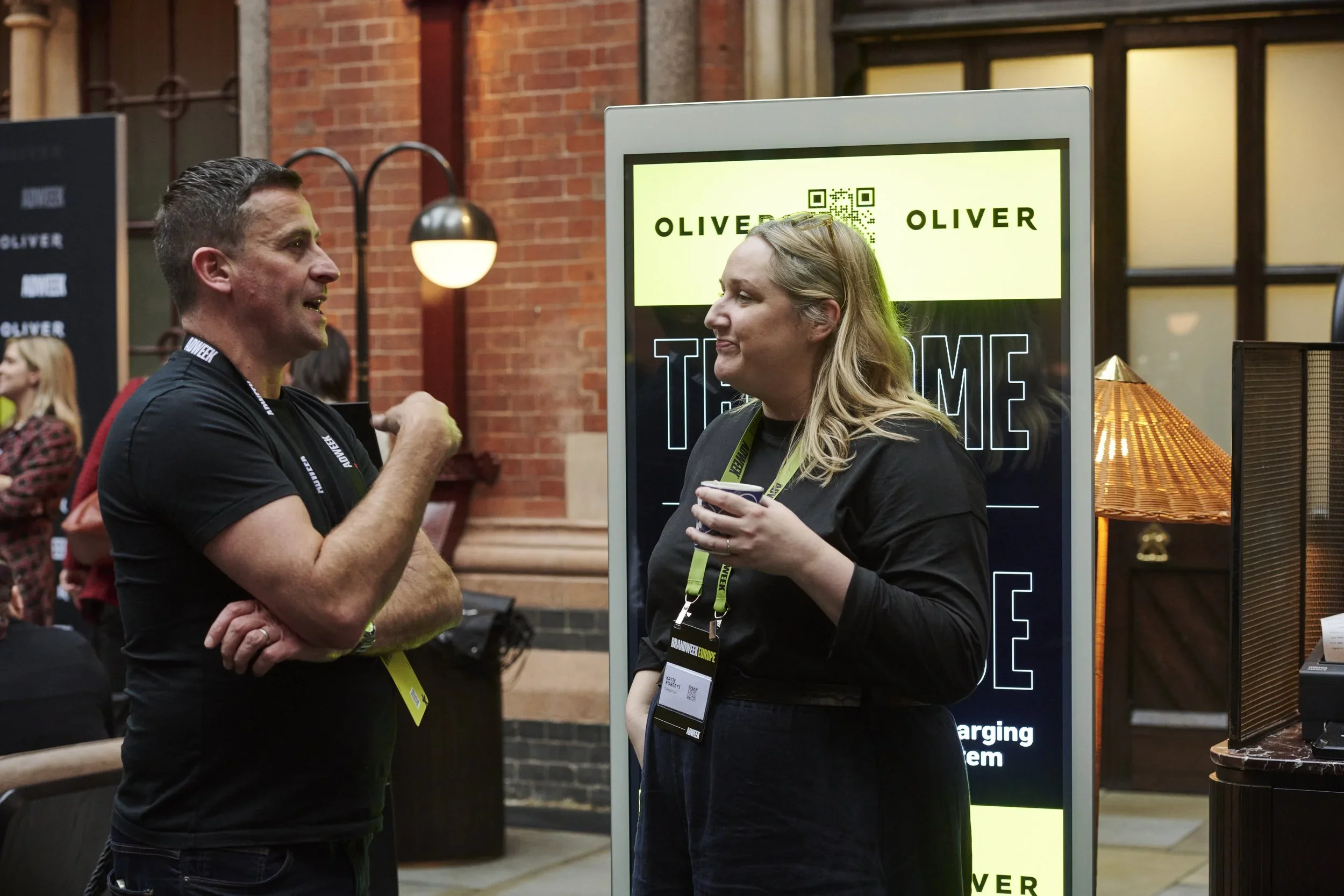 Two people are having a conversation indoors at a conference or event, with a digital sign behind them displaying the name 'Oliver' and a QR code. The man on the left is gesturing with his right hand, and the woman on the right is holding a cup and listening. Both are wearing name badges and lanyards.