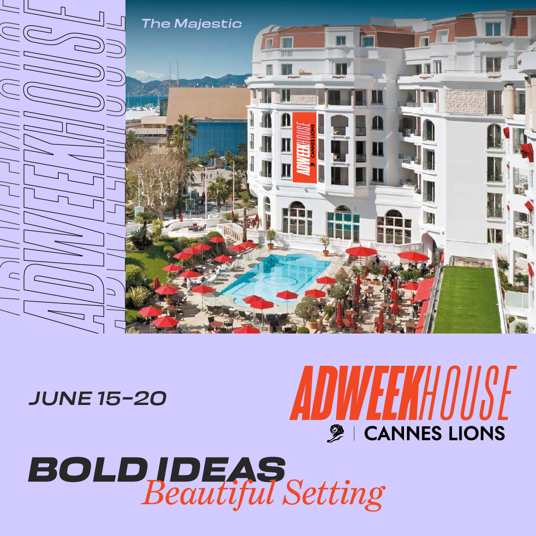 A luxury hotel with a large white building featuring multiple balconies and red awnings. There is a pool area with red umbrellas and lounge chairs, situated among palm trees and greenery. The background includes a view of mountains and water, suggest