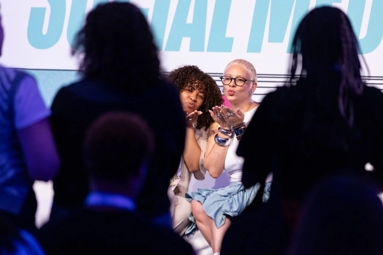 Two women blow kisses towards a group of people at a conference or event, with a large sign behind them.