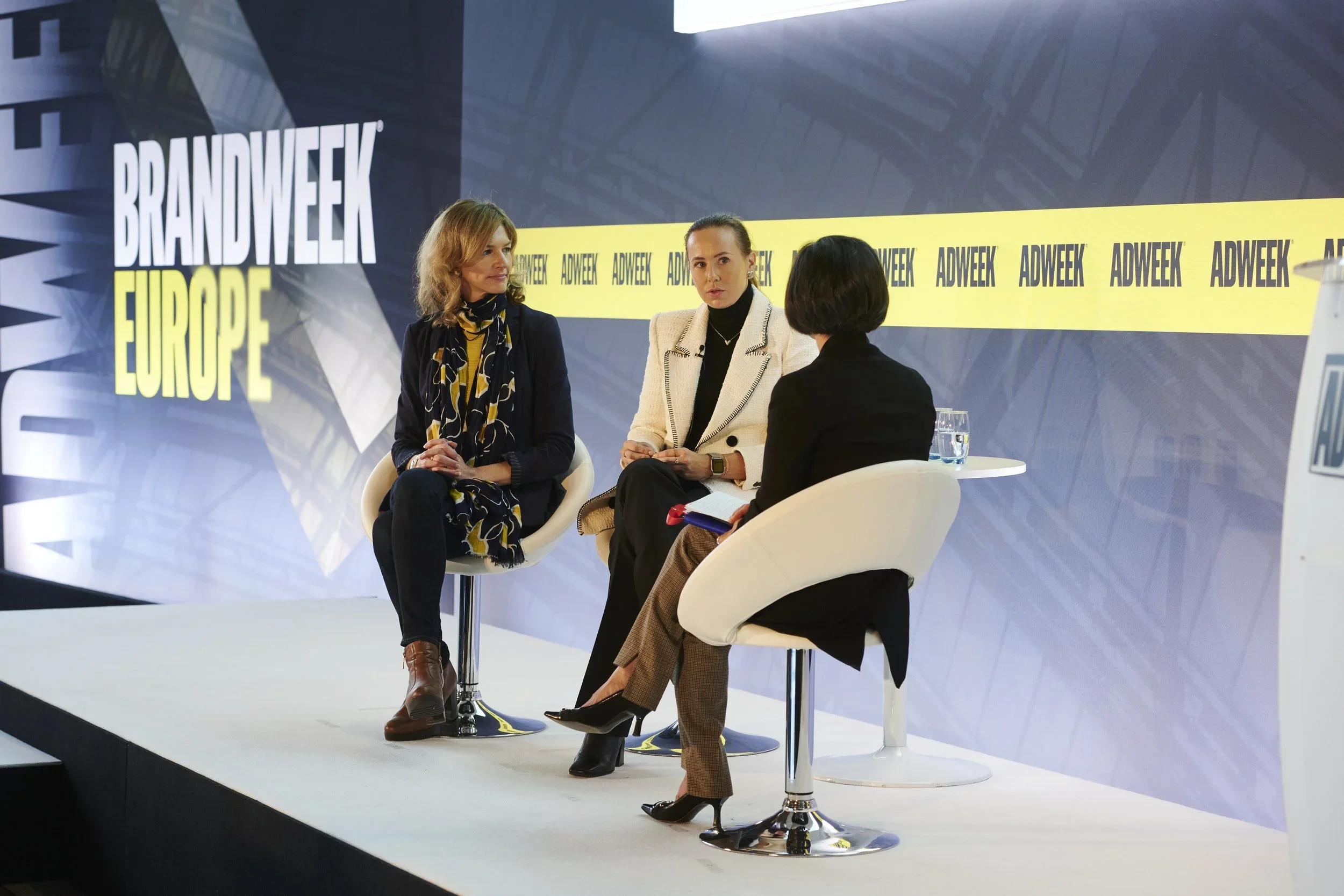 Three women sitting on stage during a panel discussion at a conference. The large background screen displays the words "BRANDWEEK EUROPE" and "ADWEEK" repeatedly.