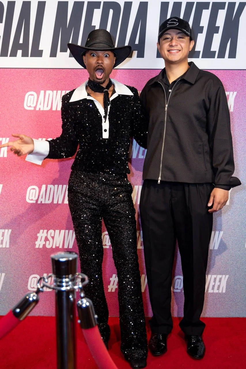 A person dressed in a black shiny sequined outfit with a wide-brimmed hat and expressive facial expression stands next to a person in casual black sportswear, both posing on a red carpet at an event with a pink and gray backdrop.