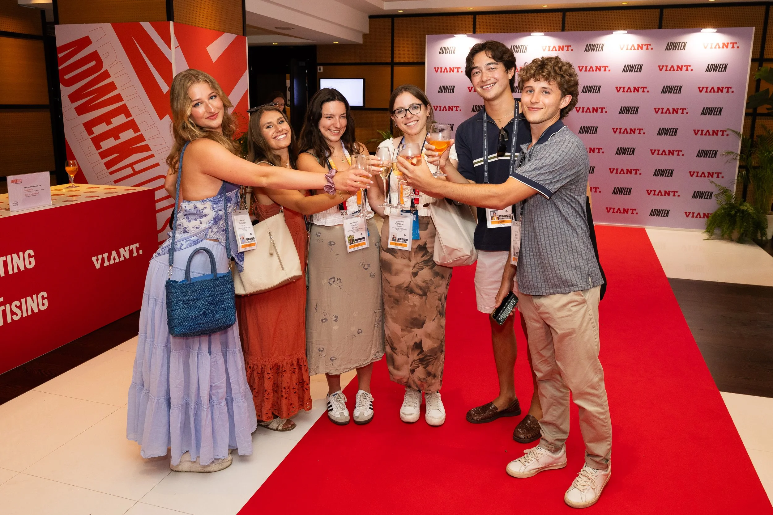 Group of young people celebrating with drinks at a conference or event, standing on a red carpet in front of a backdrop.