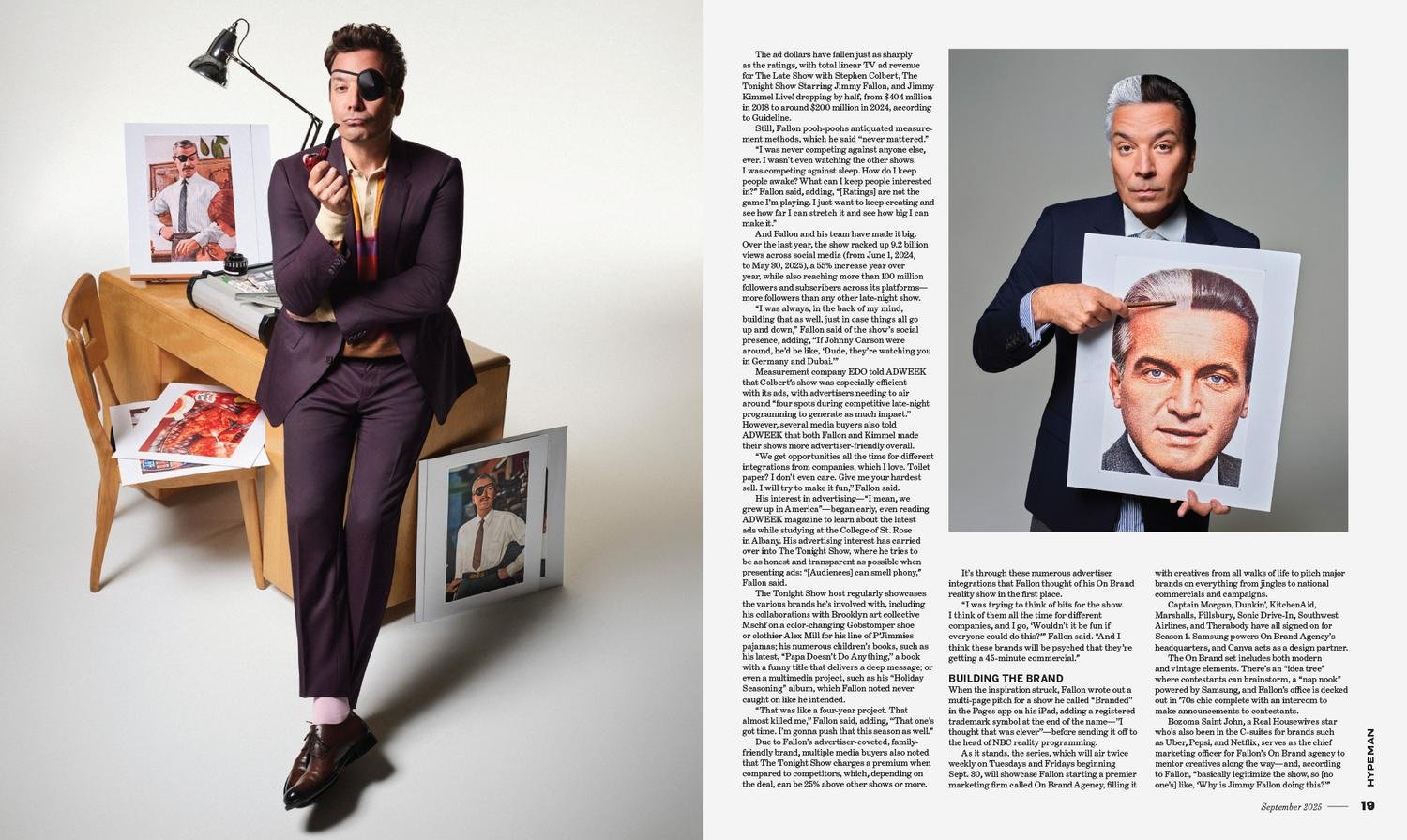 A man dressed in a dark suit and wearing a black eyepatch is sitting on a wooden chair, holding a pipe to his mouth with one hand and resting his other arm on a desk. There are magazine photographs of himself on the desk and leaning against it, and a