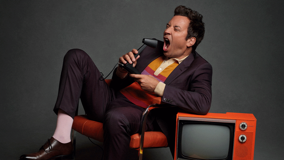 Jimmy Fallon in a suit sitting on an orange chair, singing into a vintage microphone, with an old orange television to his right.
