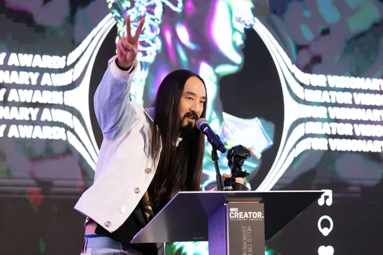 Steve Aoki speaking at a podium during an award ceremony, making a peace sign with his right hand, with a large screen displaying colorful graphics and partially visible text in the background.