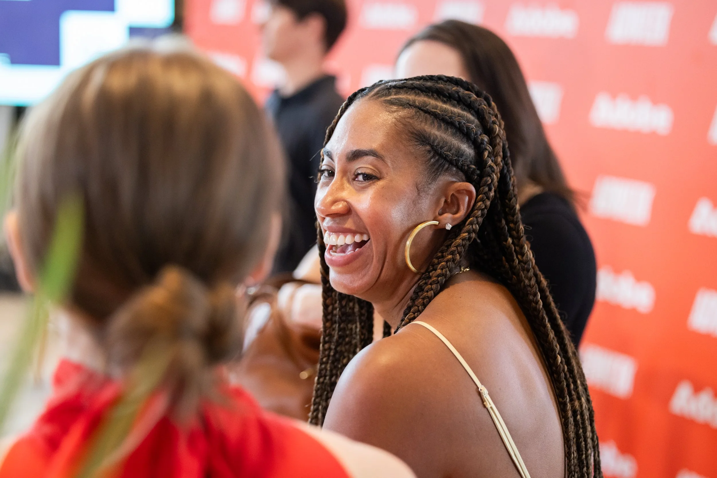 A woman with braids smiling and talking to another person in front of a red background with white text.