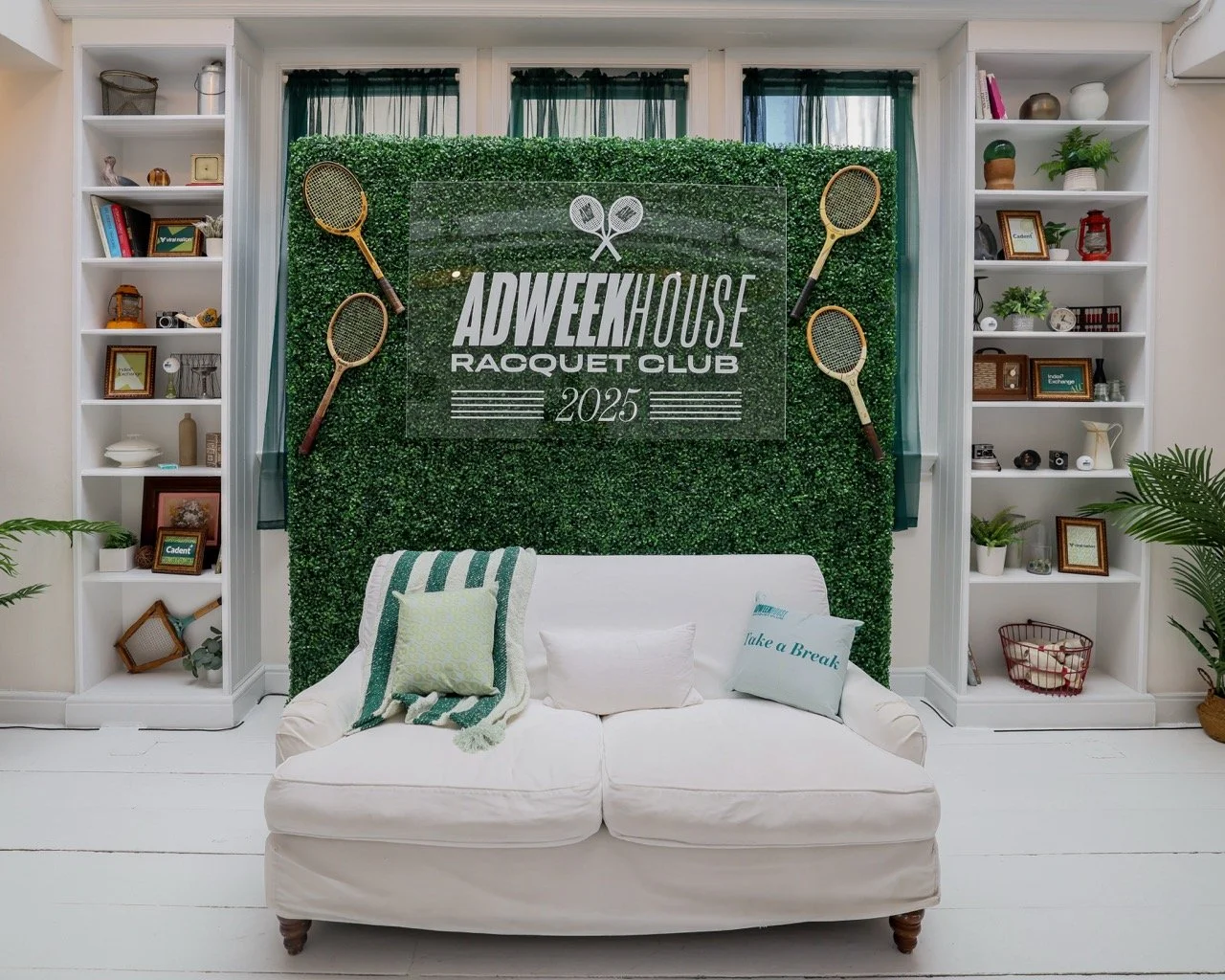 Indoor wall setup with green artificial hedge backdrop, event banner reading 'ADWEEK HOUSE RACQUET CLUB 2025', four tennis rackets mounted on the hedge, white couch with pillows, surrounded by white bookshelves with decorative items, plants, and phot