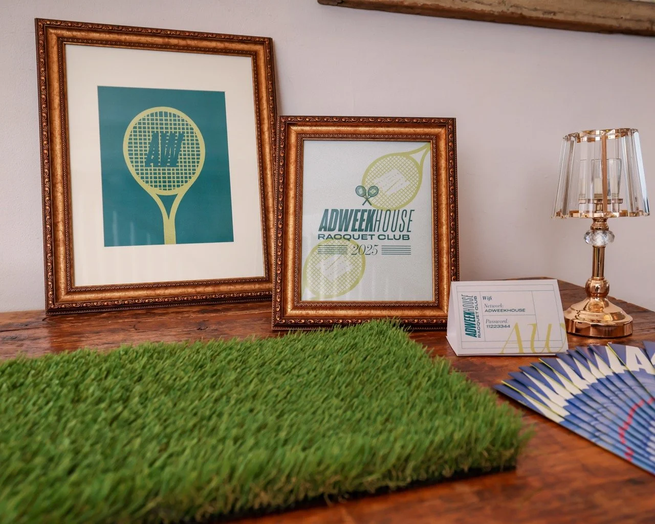 Framed posters of tennis rackets and a raffle event display on a wooden table with a small lamp, a green artificial grass mat, and informational flyers.