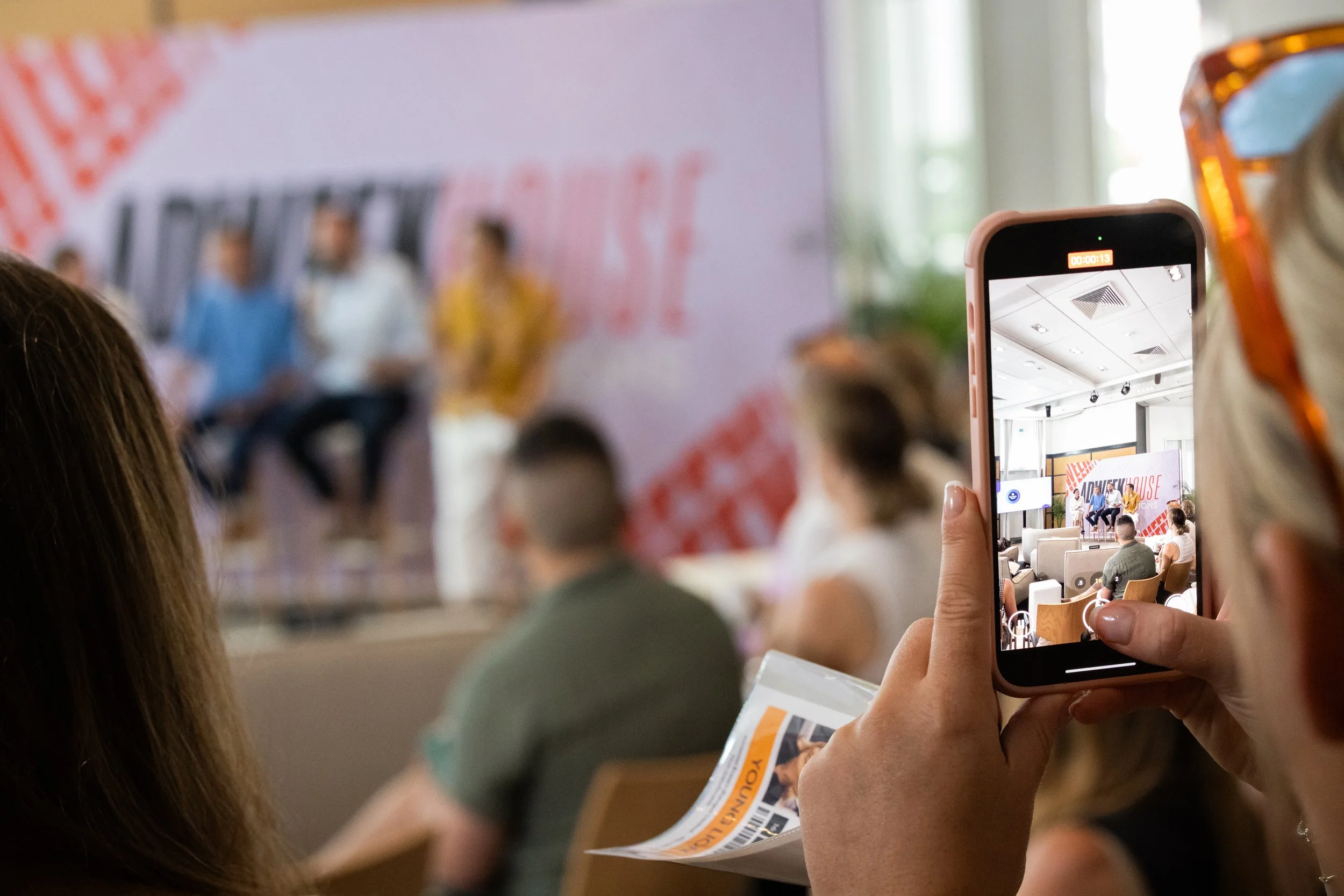 Person recording a panel discussion on smartphone during conference with blurred audience in background.