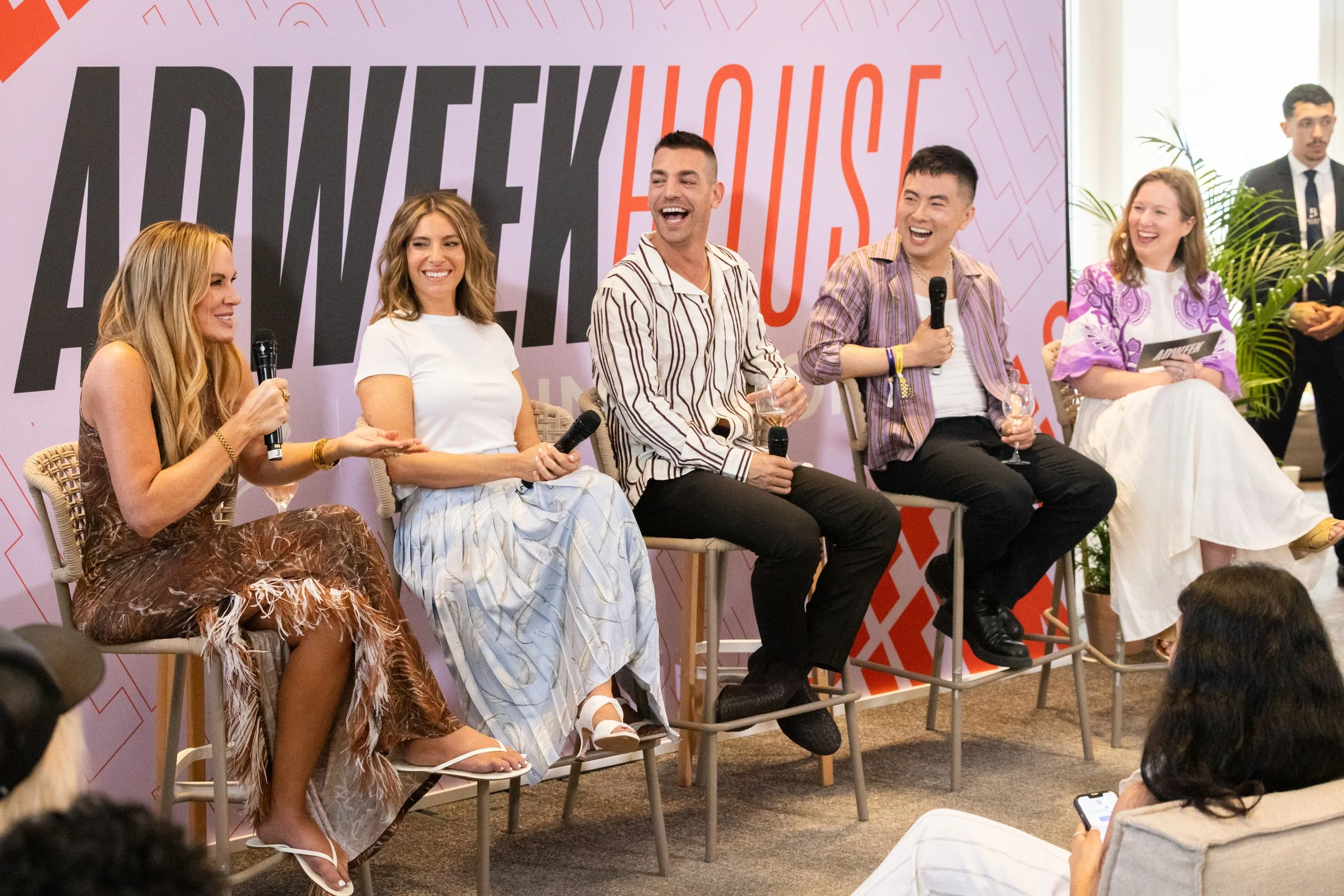 A panel of five people sitting on stage with microphones, smiling and engaging in a discussion at an event with a large purple and red backdrop that reads "ADWEEK HOUSE."