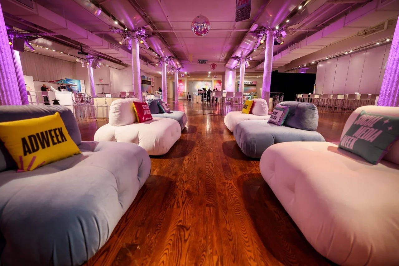 An indoor event space with velvet couches and colorful pillows, purple lighting, wooden floors, and a disco ball hanging from the ceiling.
