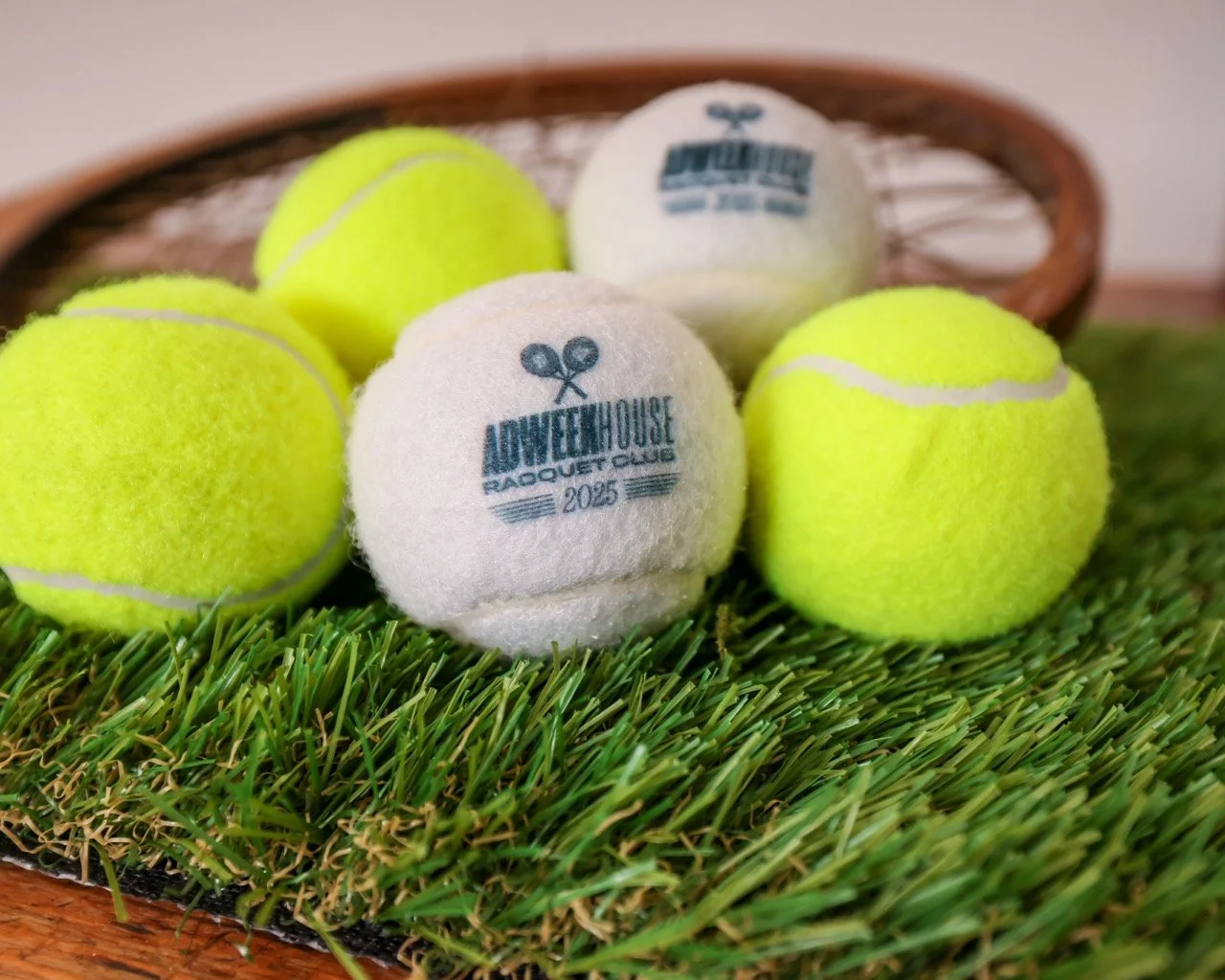 Six tennis balls, two white and four yellow, with the white ones displaying a logo and the word 'Adventure House' and a year, placed on artificial grass in front of a wicker basket.