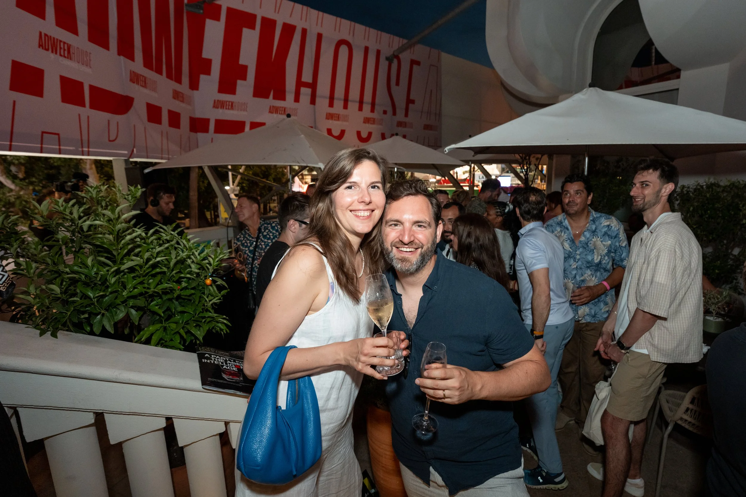 Two people, a woman in a white dress holding a glass of wine and a man in a navy shirt with a glass of wine, smiling at a social gathering or party with a crowd of people in the background, outdoor setting with umbrellas and greenery.