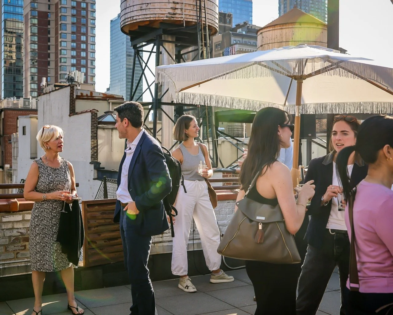 People socializing on a rooftop in an urban area with modern skyscrapers, a water tower, and outdoor umbrellas.