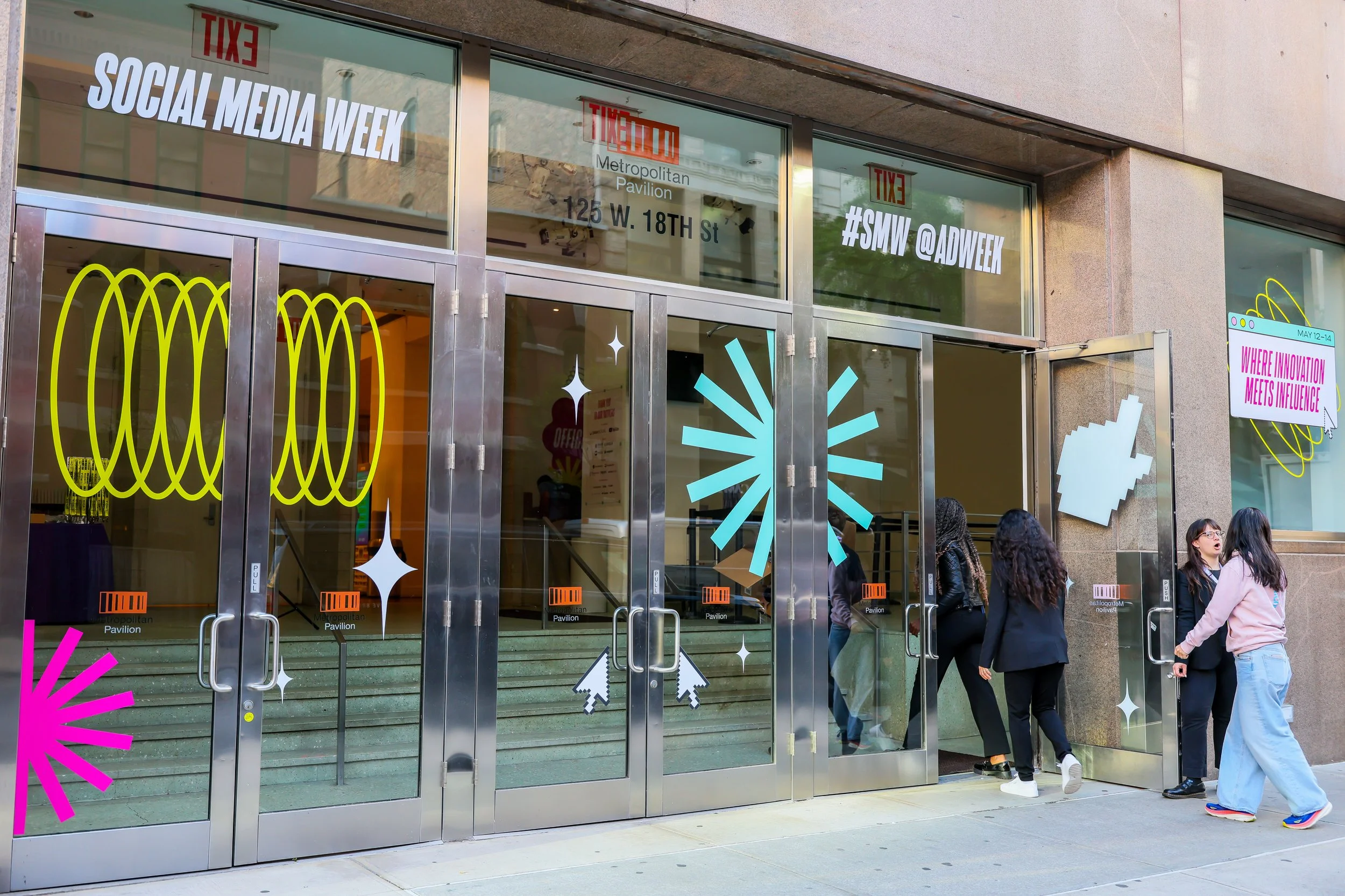 Glass doors decorated with colorful neon-style graphics and signs, with a group of people entering the building, which is identified as the Metropolitan Pavilion, hosting Social Media Week, with signs indicating the event's social media hashtags.