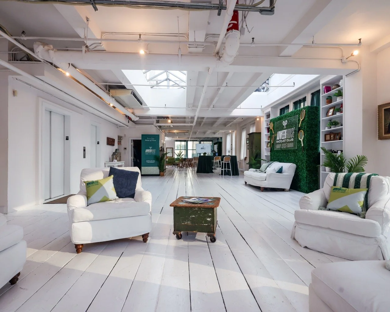 Spacious lounge area with white sofas, wooden coffee table on wheels, decorative pillows, books, and indoor plants, with a green hedge wall featuring tennis-related signage and multiple shelves in the background.