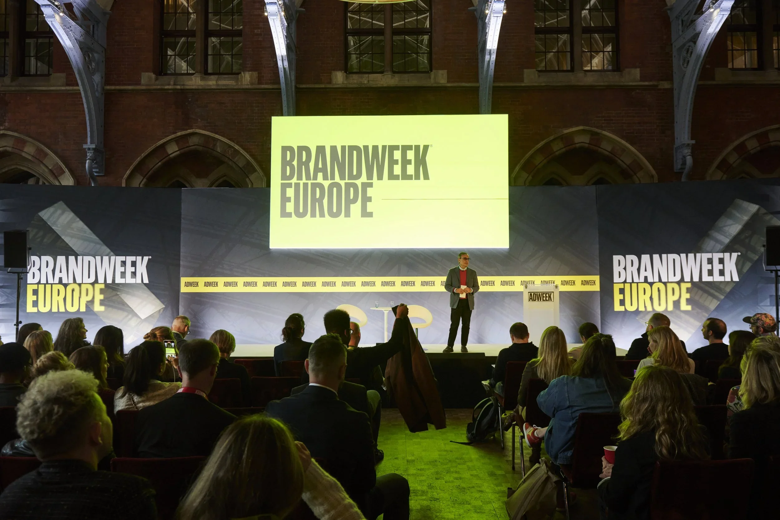 Speaker on stage at Brandweek Europe conference with audience seated in front, large screens displaying 'BRANDWEEK EUROPE', in an industrial-style venue with exposed brick walls and metal beams.