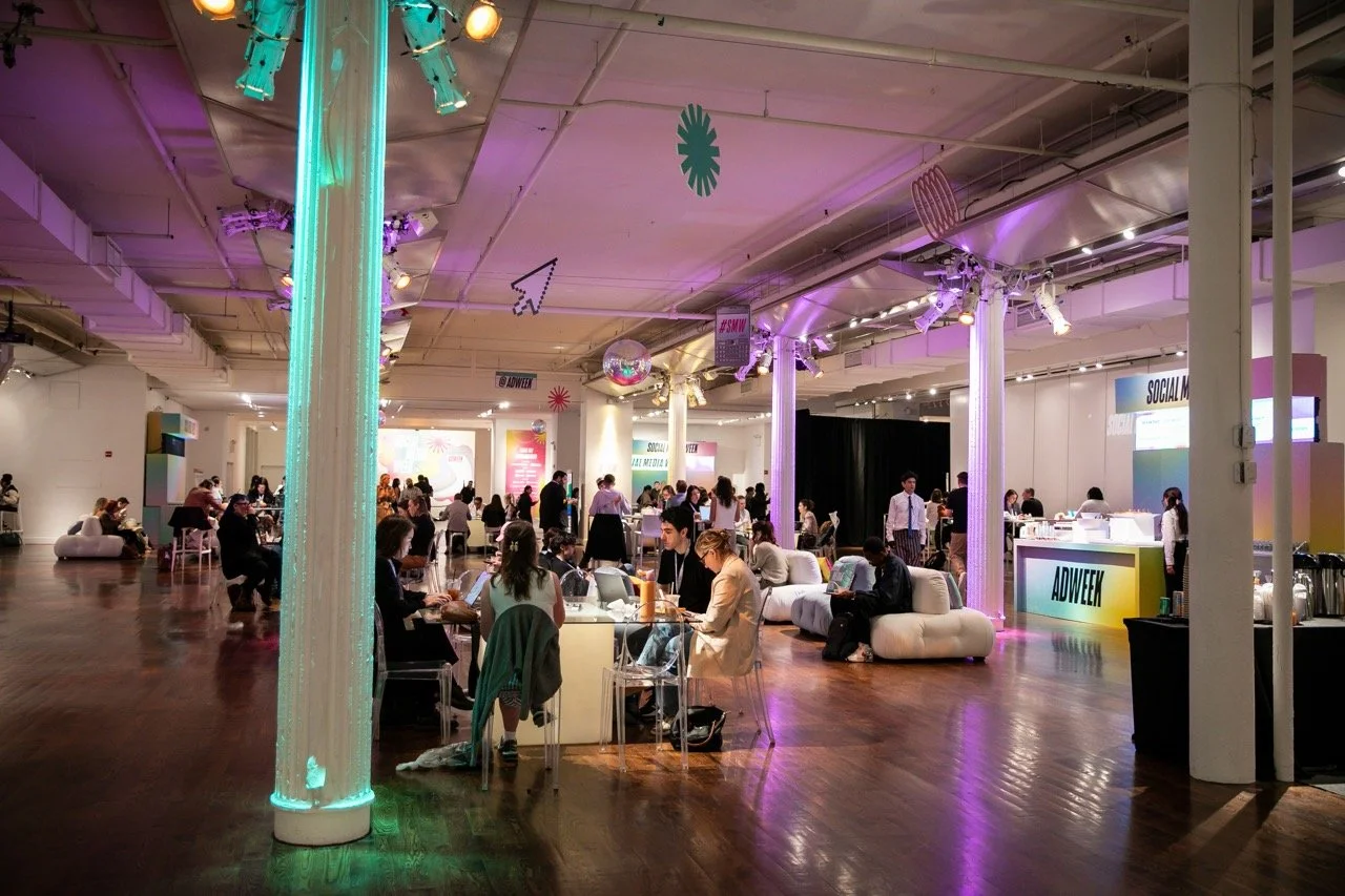 Indoor conference or event space with people sitting at tables, standing, and walking. The room is decorated with purple and teal lighting, hanging decor, and a disco ball. There are signs with the names 'ADWEEK' and 'SOCIAL' and equipment such as screens and a bar area.